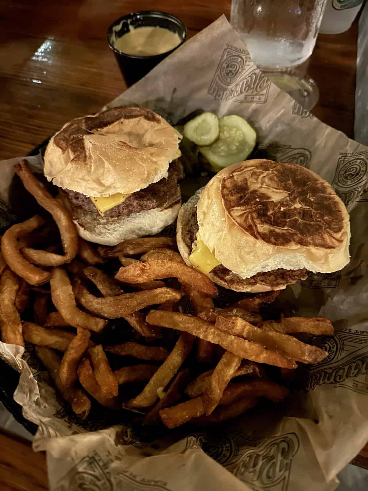 the_pharmacy_nashville_burger - Adventures with Carli Two cheeseburgers with melted cheese and toasted buns, accompanied by fries, pickle slices, and a cup of sauce on a wooden table.