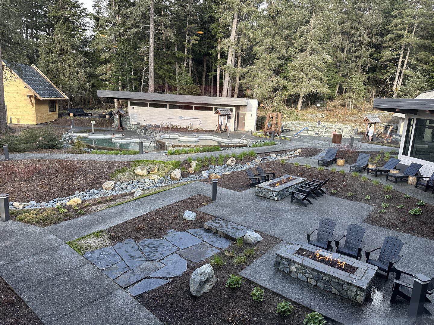 Outdoor spa area with stone pools, wooden structures, and numerous black lounge chairs set against a forest backdrop of tall trees and evergreens.