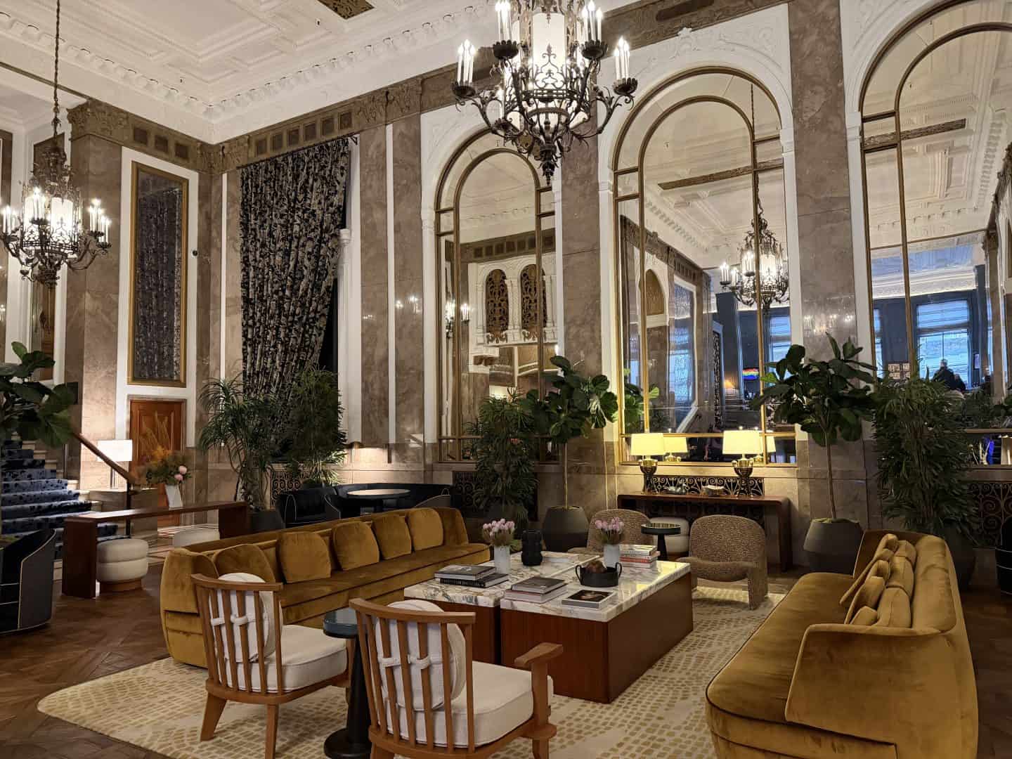 Luxurious hotel lobby with marble walls, chandeliers, and mustard-yellow sofas.