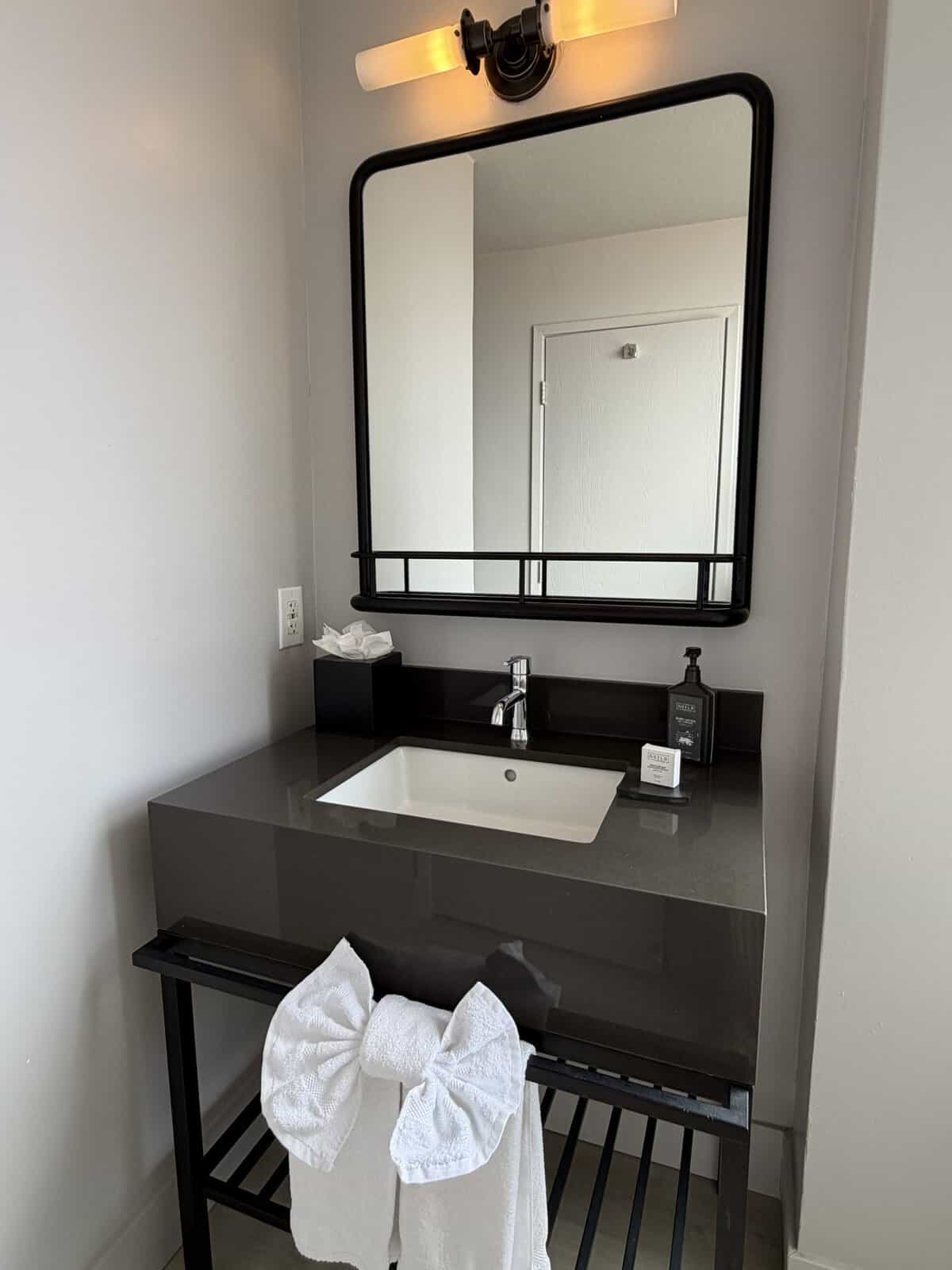 hotel_garrett_sf_bathroom - Adventures with Carli Modern bathroom vanity with dark countertop, white sink, chrome faucet, and a large black-framed mirror above it.