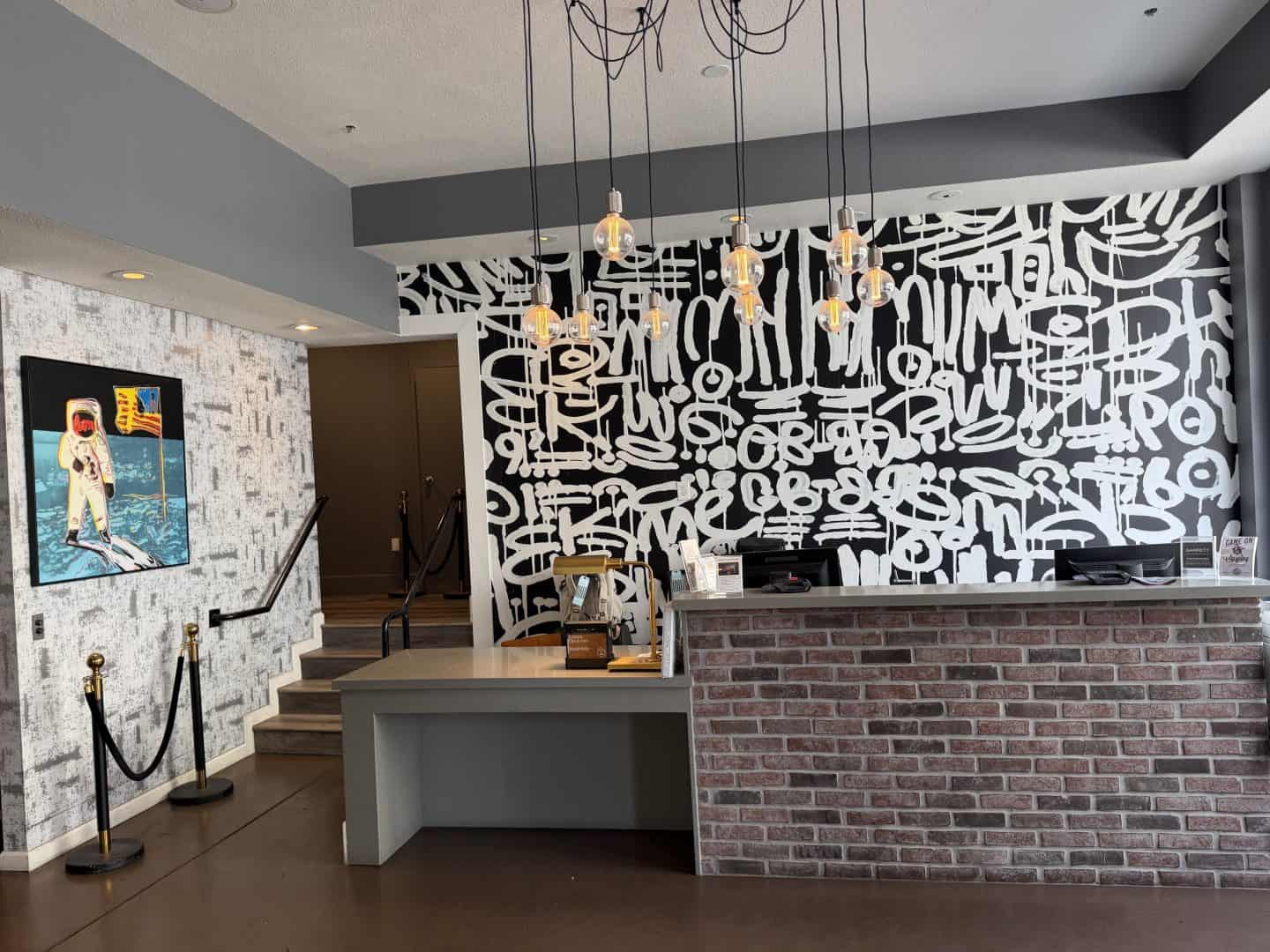 hotel_garrett_sf_reception - Adventures with Carli Interior lobby with hanging Edison bulb lights above a front desk and a black-and-white abstract mural on the wall behind. A short staircase is to the left, brick counter to the right.