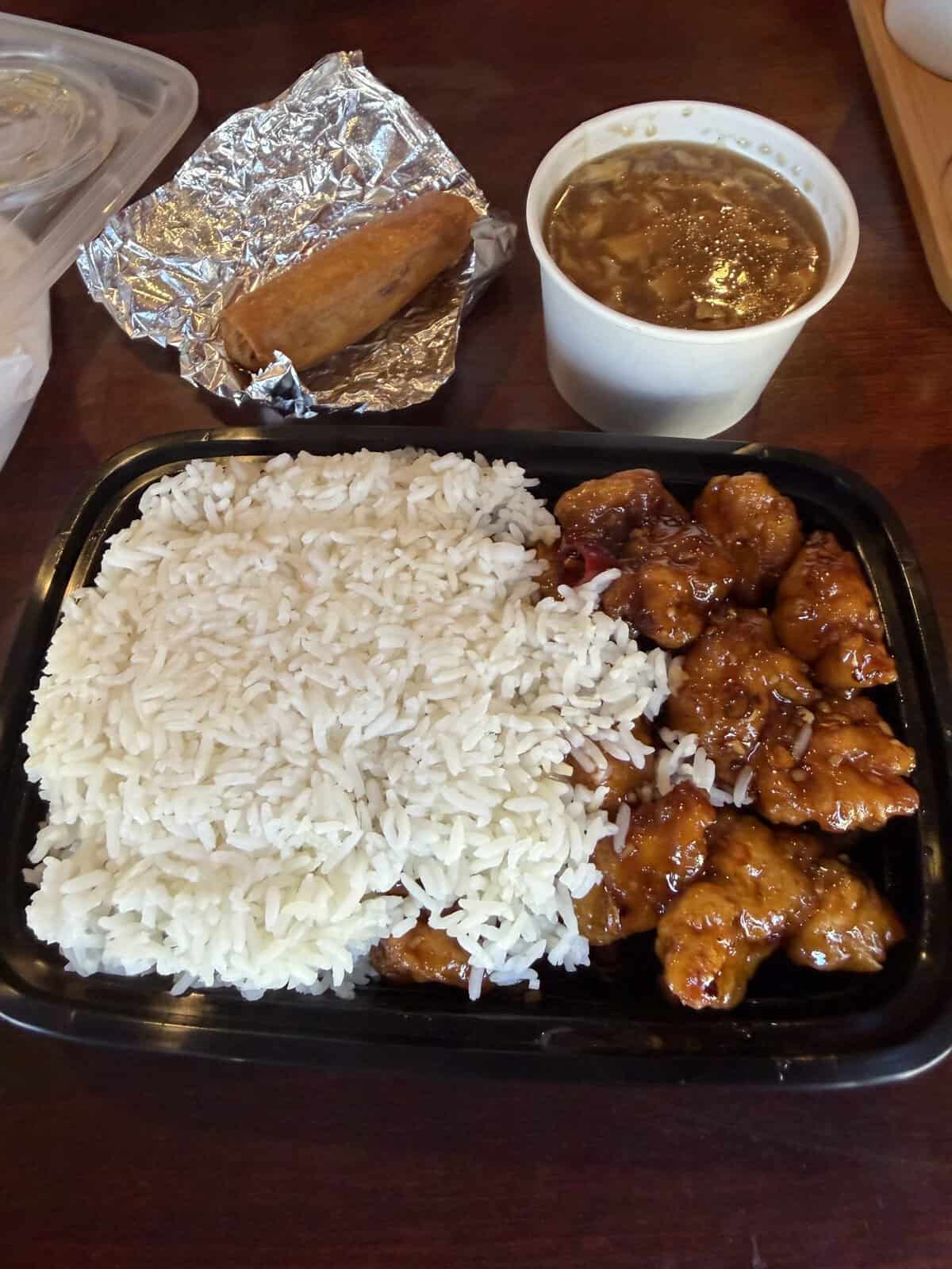 large plate of general tso chicken and small cup of broth soup and eggroll on tin foil