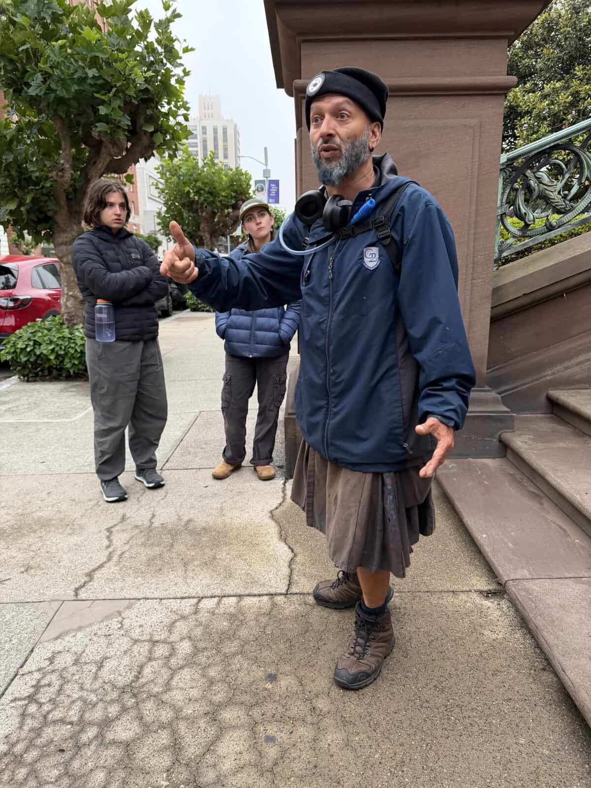 San Fran Ghost Tour guide with a gray beard in a black beanie and blue jacket shares story while two young people stand behind him on a city sidewalk listening attentively.