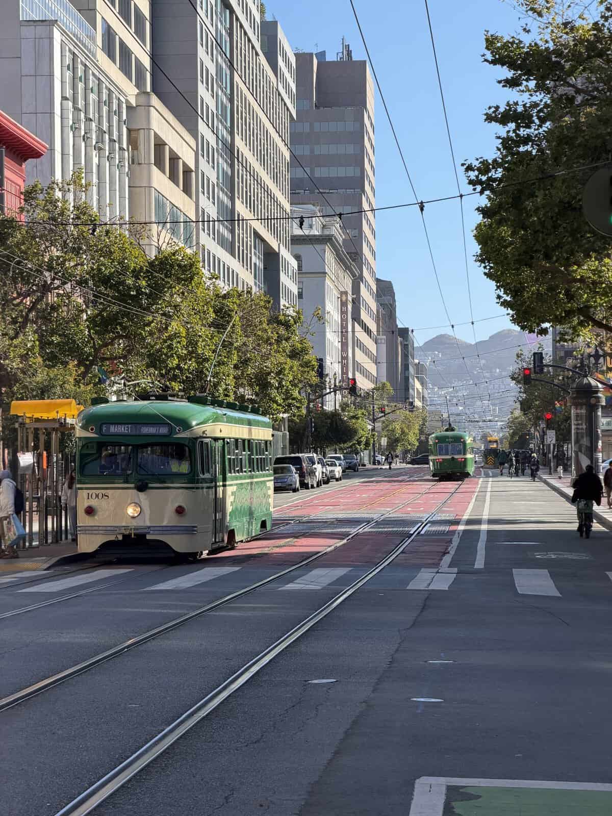 sf_market_streetcar - Adventures with Carli Green and white vintage trolleybus on a city street with tram tracks, tall buildings lining the avenue and trees against a clear blue sky.