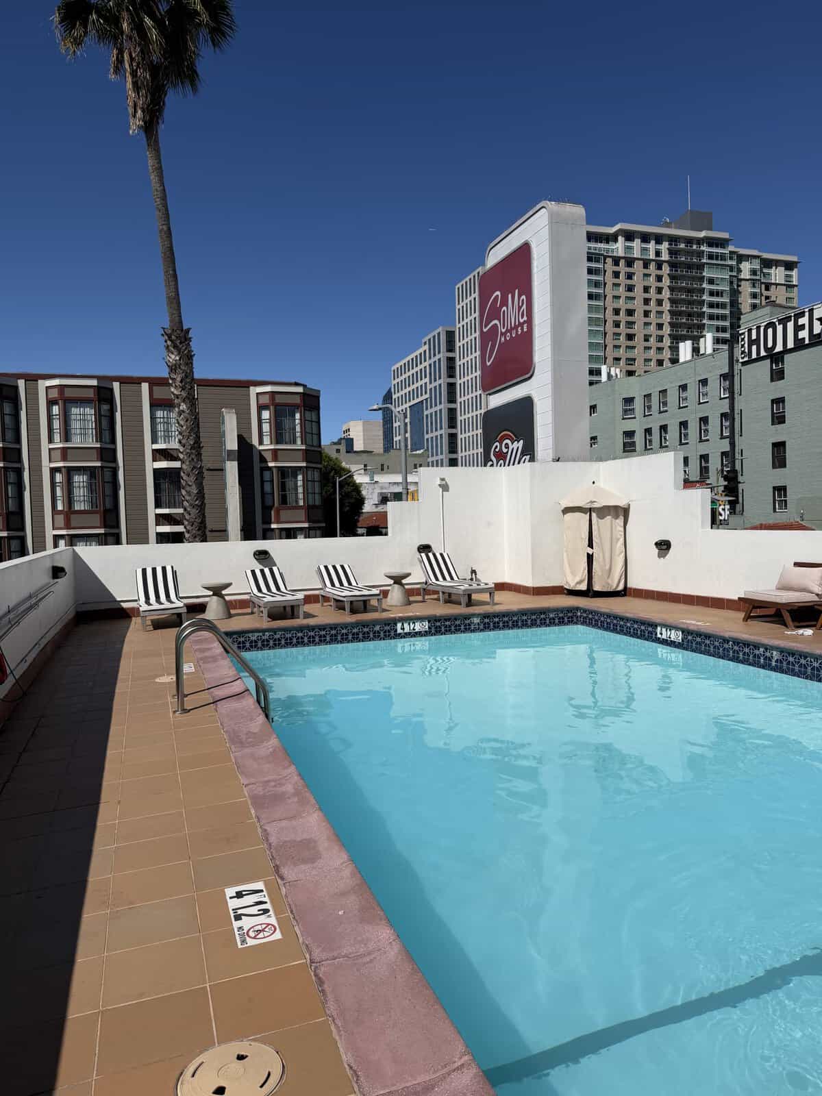 soma_house_pool - Adventures with Carli Rooftop hotel pool with striped lounge chairs, palm tree, and a clear blue sky against a city skyline.