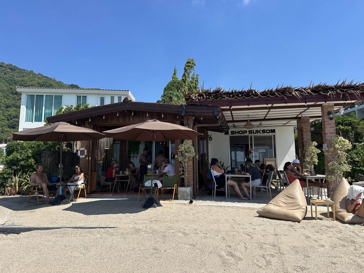 sun_chasers_cafe_haad_rin - Adventures with Carli People dining at an outdoor cafe under brown umbrellas, in front of a white building and clear blue sky.