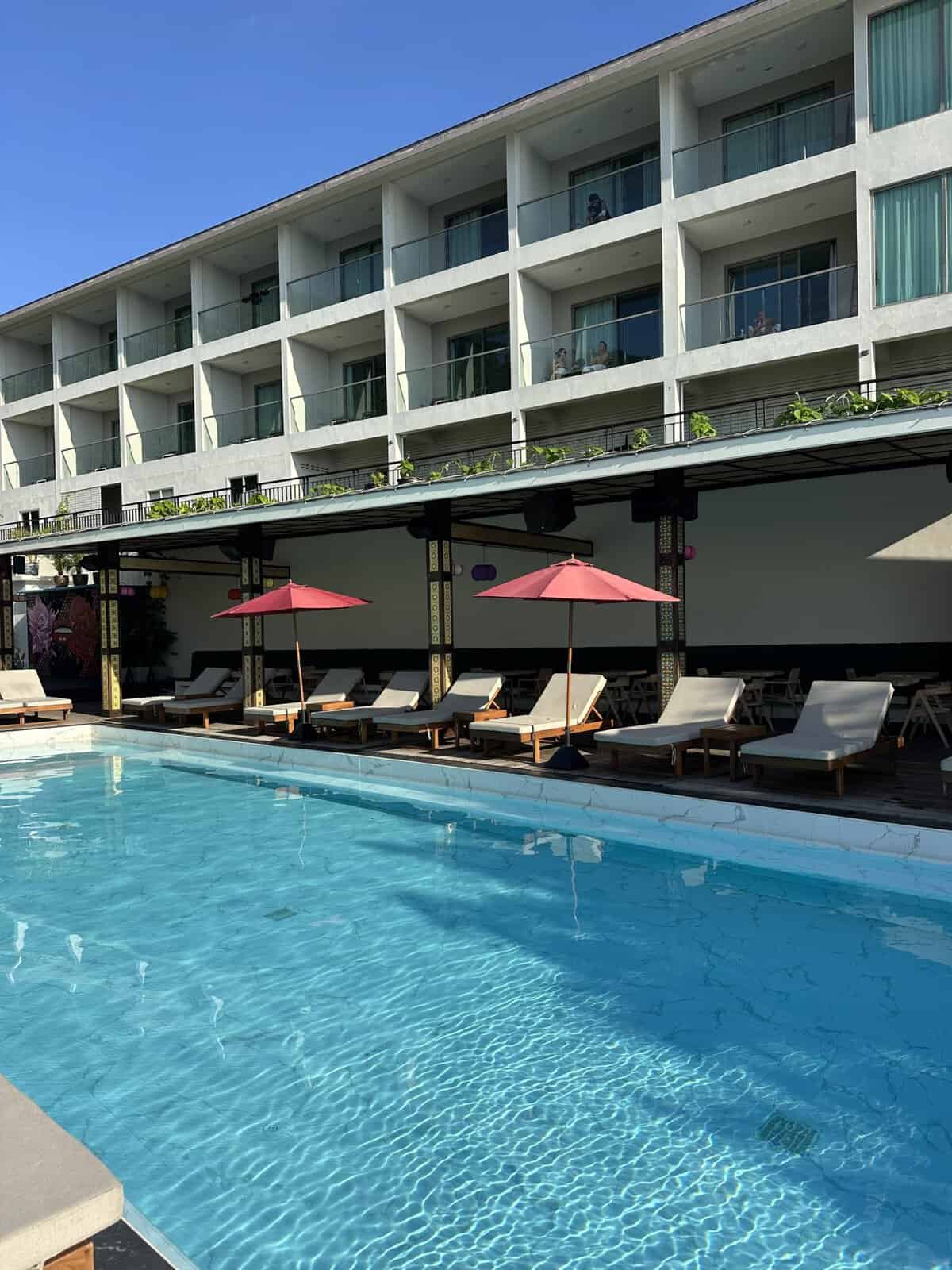 sunset_beach_club_hotel_pool - Adventures with Carli Poolside view of a modern hotel with multiple glass balconies facing the pool area and sun loungers under pink umbrellas.