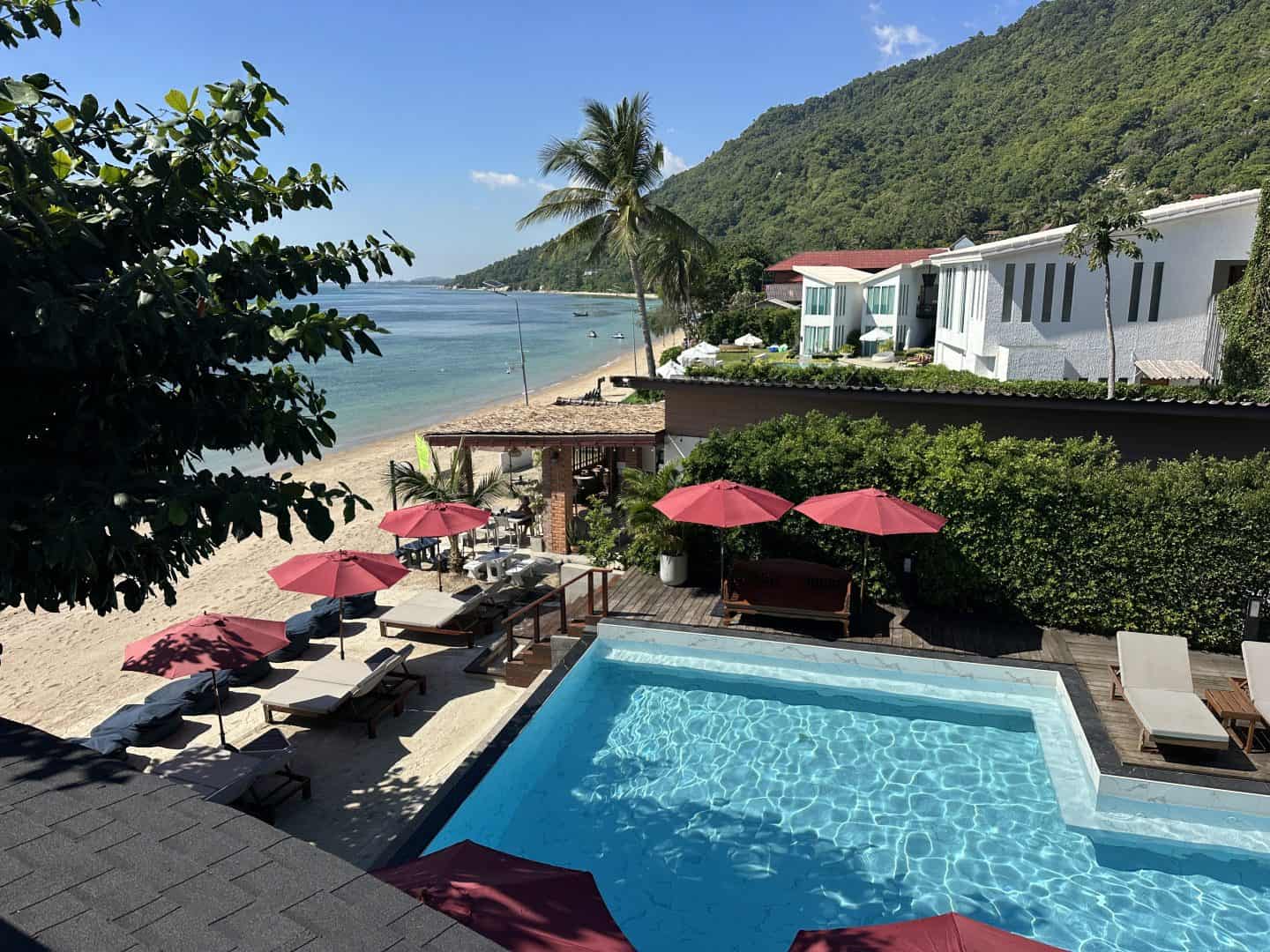 sunset_beach_club_hotel_pool_beach - Adventures with Carli Resort pool with red umbrellas and lounge chairs overlooking a sandy beach and calm sea. White buildings rise behind a green hillside.