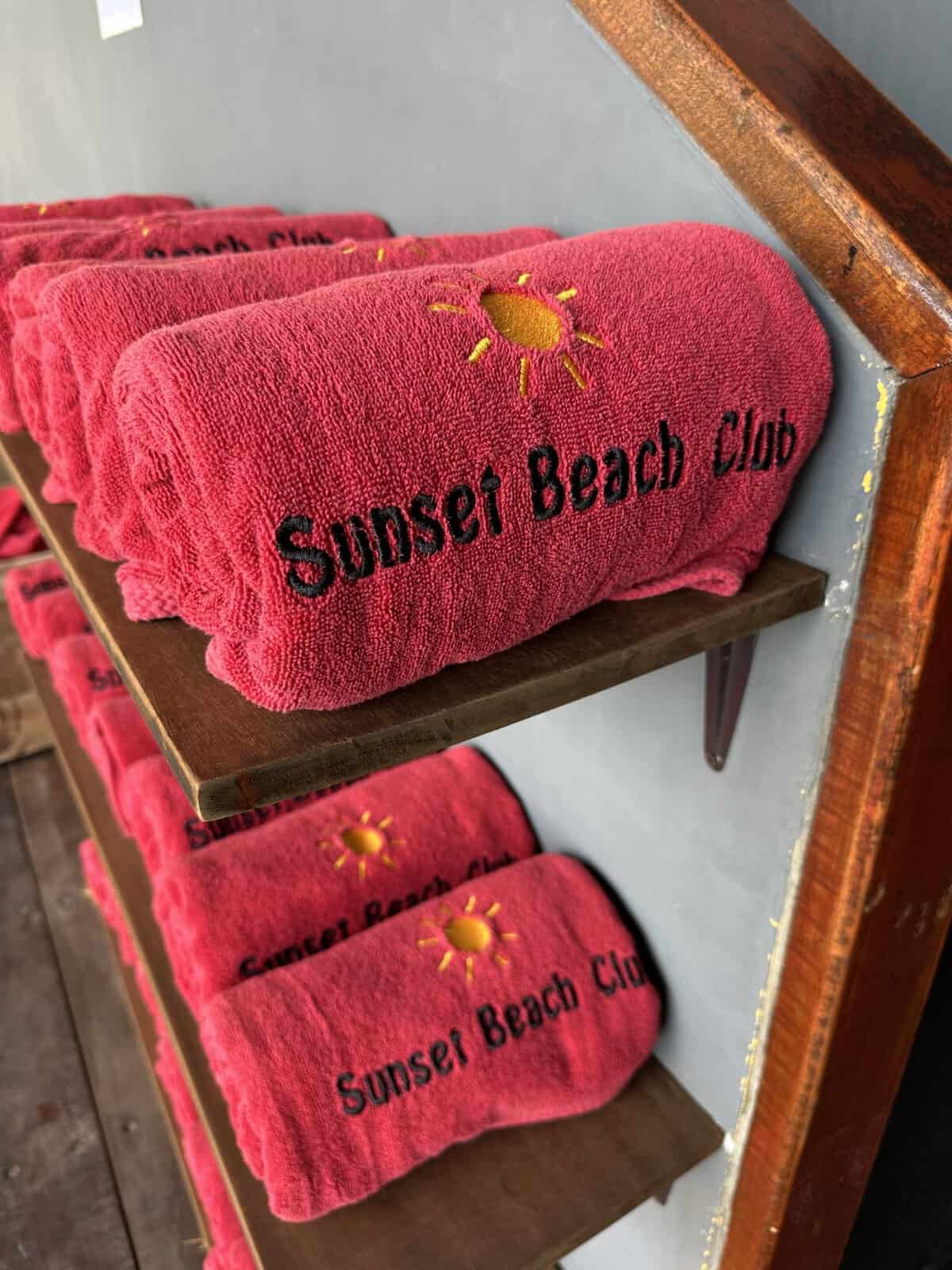 sunset_beach_club_hotel_pool_towels - Adventures with Carli Pink towel folded on a wooden shelf, embroidered with a yellow sun and Sunset Beach Club Hotel Haad Rin branding.