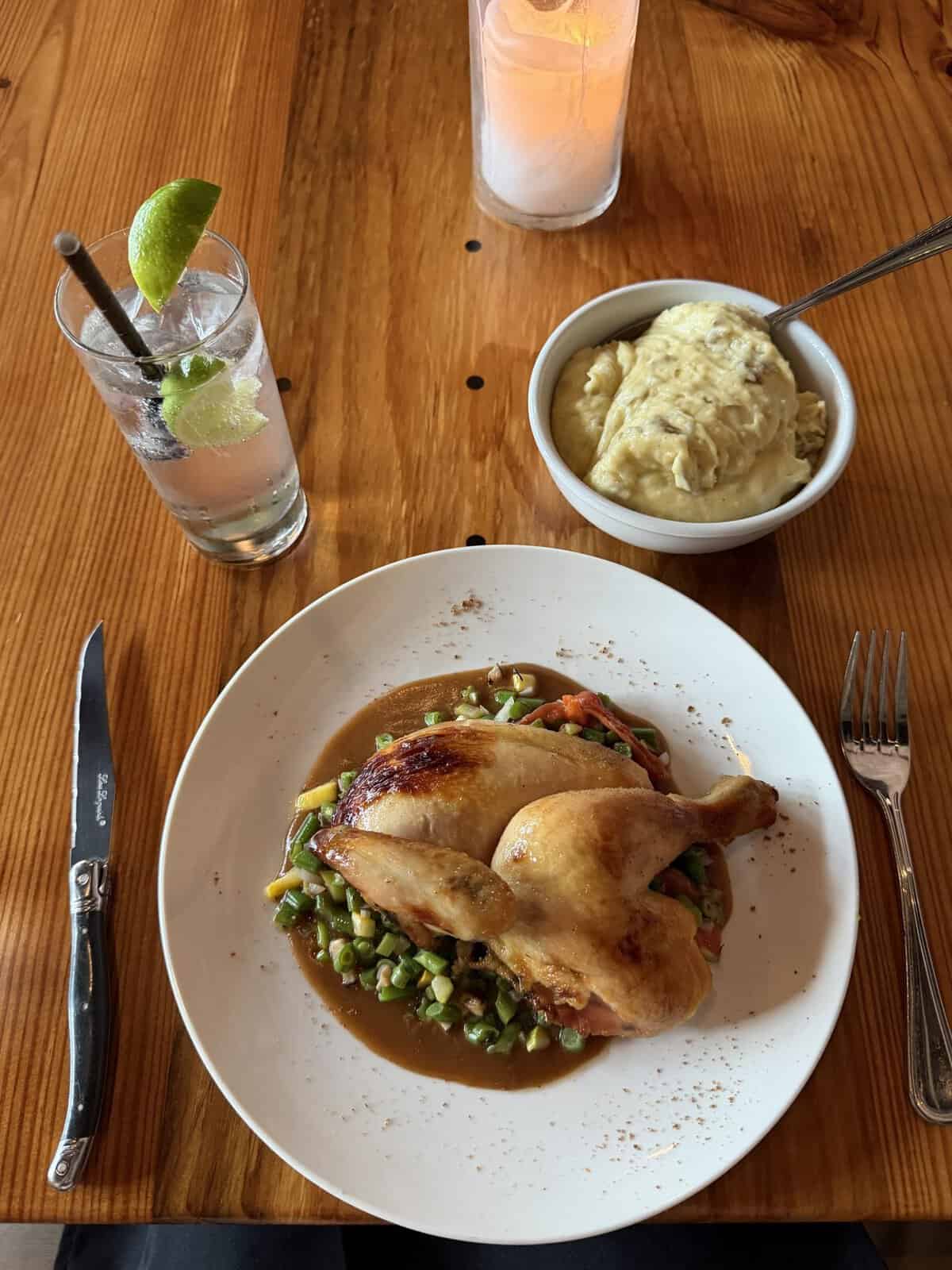 Plate with roasted chicken leg on a bed of diced vegetables in sauce, mashed potatoes in a bowl nearby on a wooden table with a fork and knife nearby.