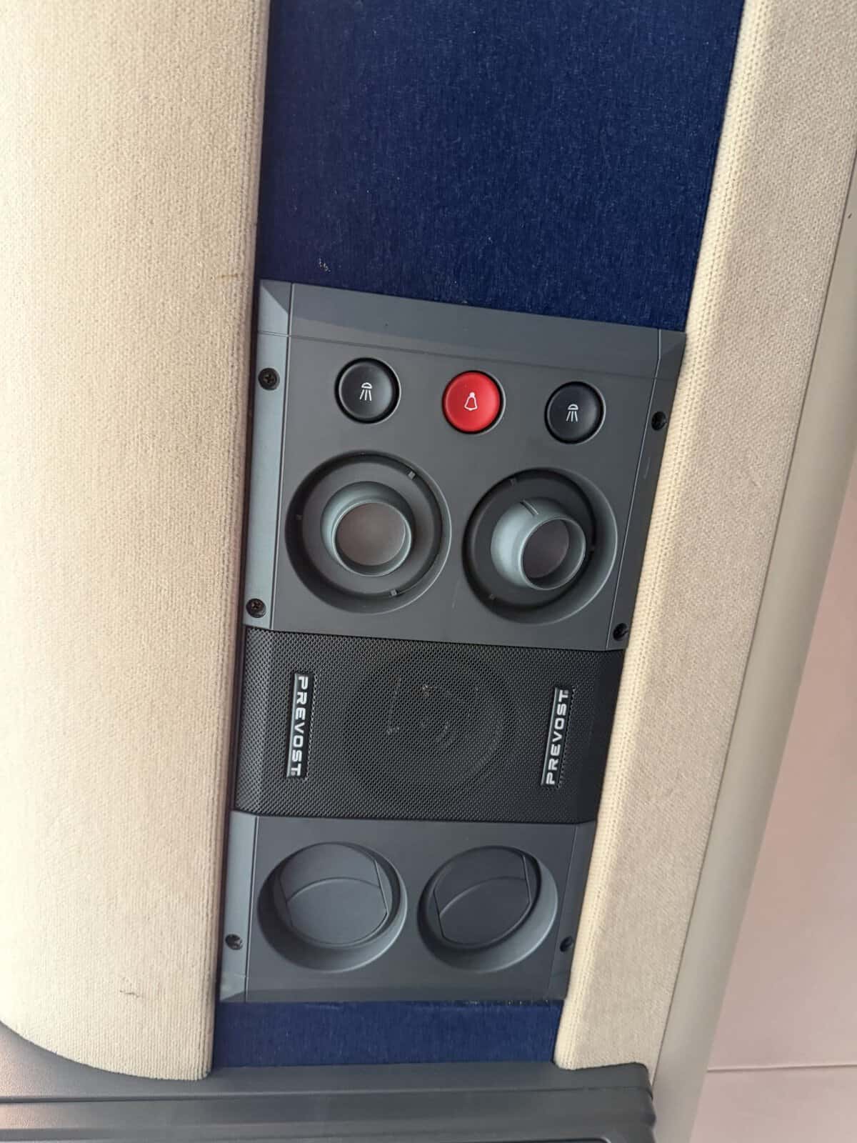 Close-up of a beige overhead control panel in a vehicle, featuring two round dials, a red center alert/bell button, and black buttons on either side, with blue fabric panels above and speaker below labeled Prevost.