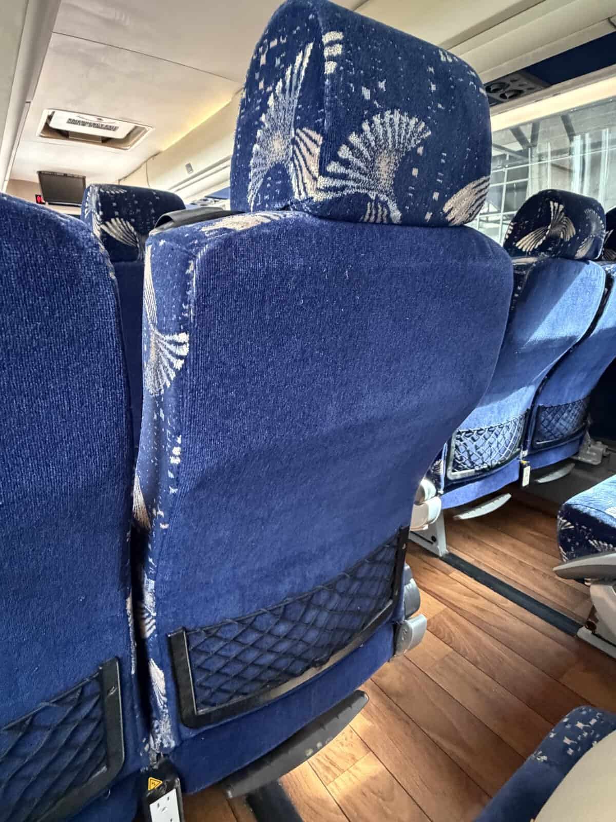 Row of blue upholstered seats with white patterns inside a vehicle, showing seat backs with mesh pockets and a wood-toned floor along an aisle.