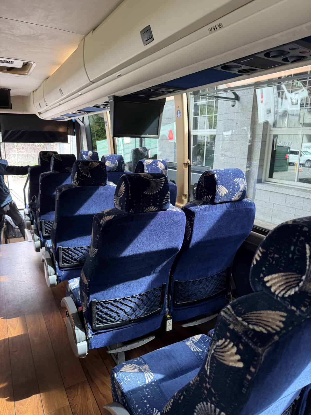 Aisle view inside a blue-seated tour bus with overhead storage and a wall-mounted TV screen.