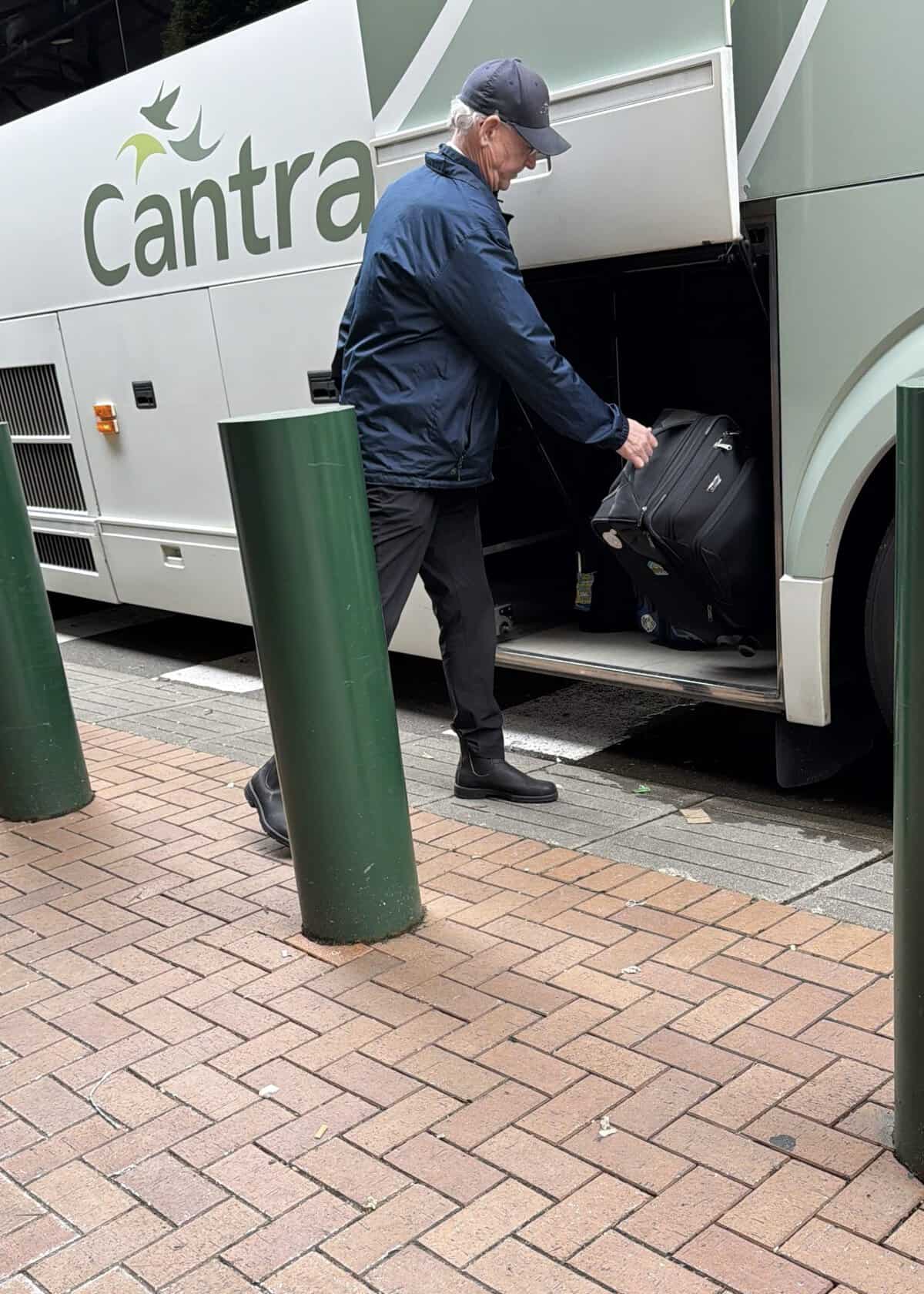 Older man in a navy jacket and cap loading a black suitcase into a large gray tour bus labeled'Cantra'
