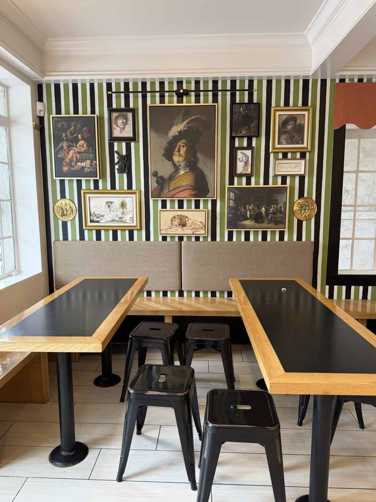 Cafe interior with a gallery-style wall of framed paintings against green, black, and white striped wallpaper; two long black-and-wood tables and metal stools.