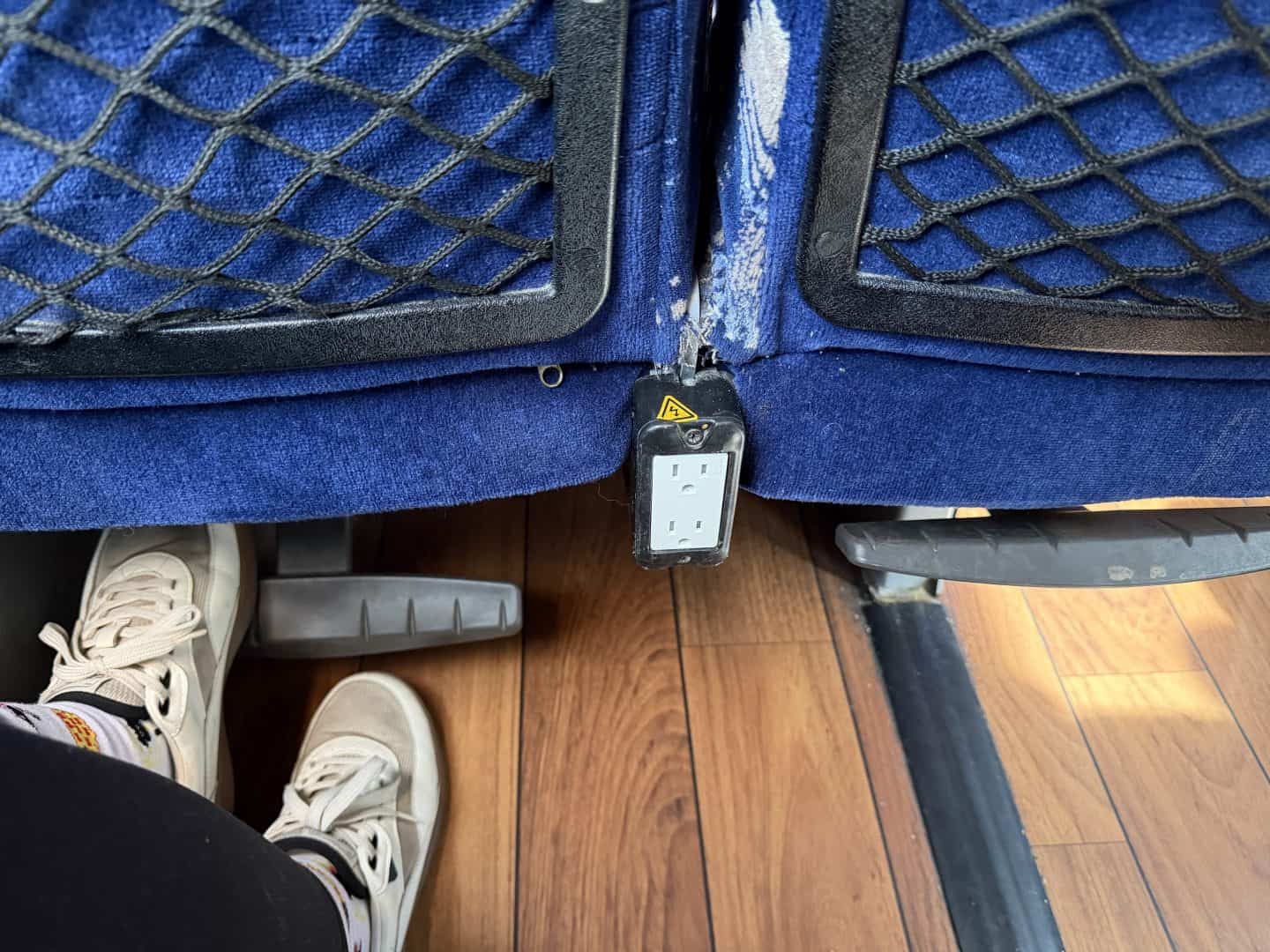 View of the underside of a blue upholstered seat with a hanging electrical outlet/strip between the seat backs, netted storage panels, and a pair of white sneakers in the foreground.
