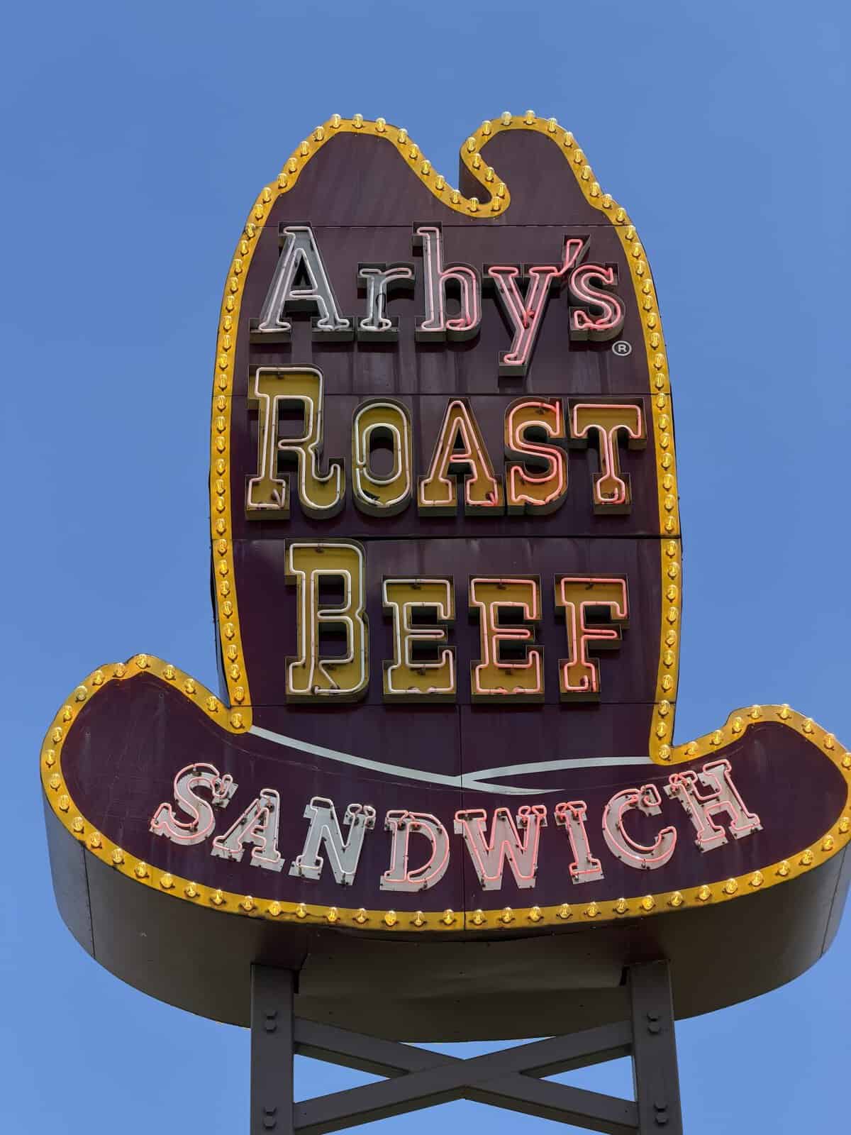 Neon Arby's Roast Beef sign on a tall maroon panel with a yellow border, set against a clear blue sky, reading 'Arby's Roast Beef' with a 'Sandwich' banner below.