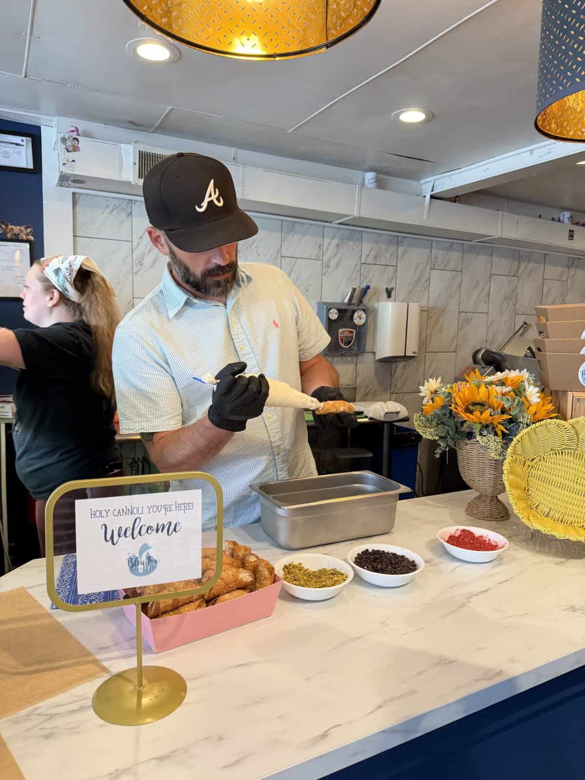 Man wearing a black cap and blue check shirt piping a cannoli at a marble counter, with bowls of toppings and a sunflower arrangement nearby.