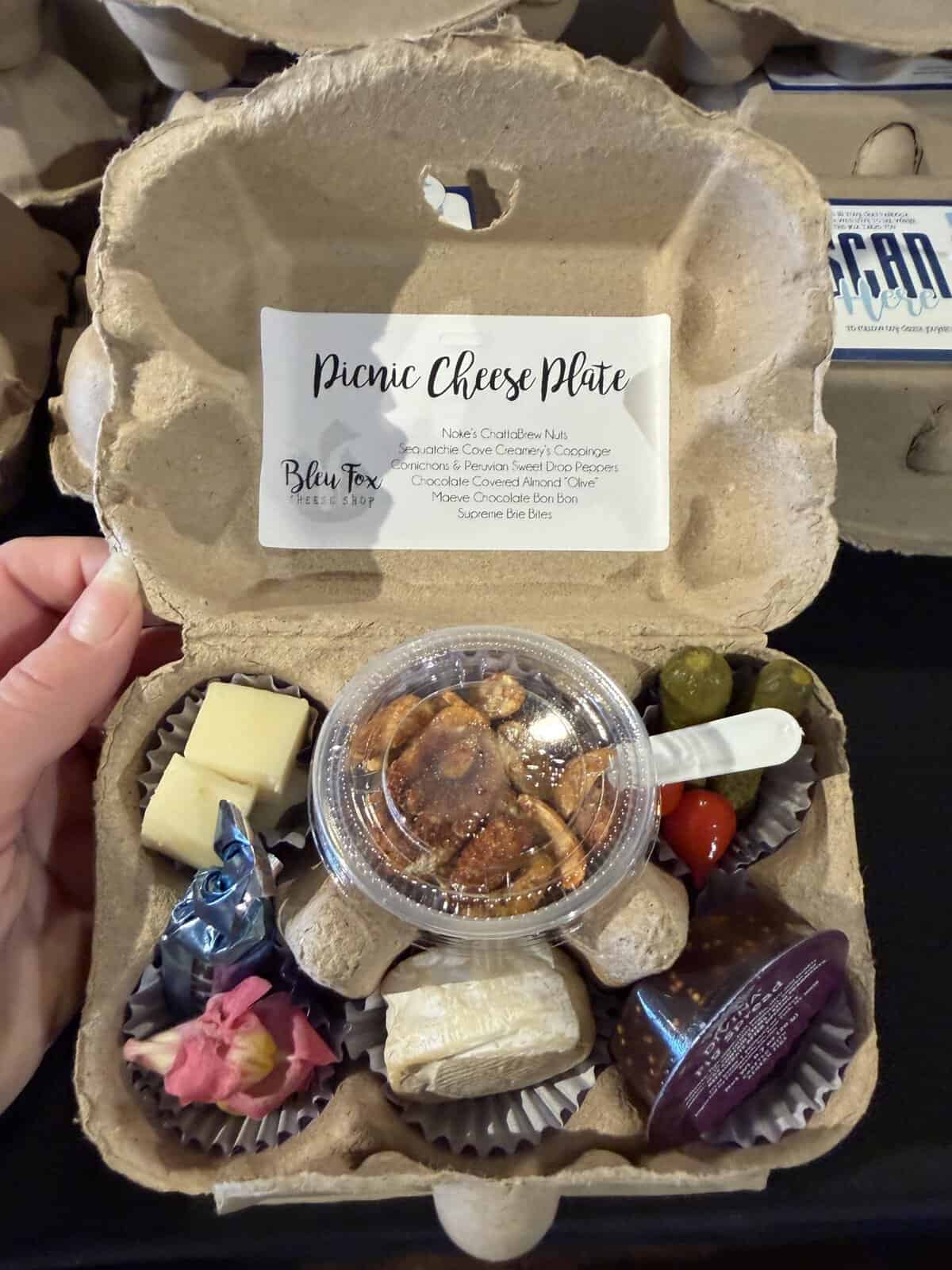 Picnic cheese plate box with cheese cubes, olives, nuts, chocolate, and wrapped treats in a cardboard tray.