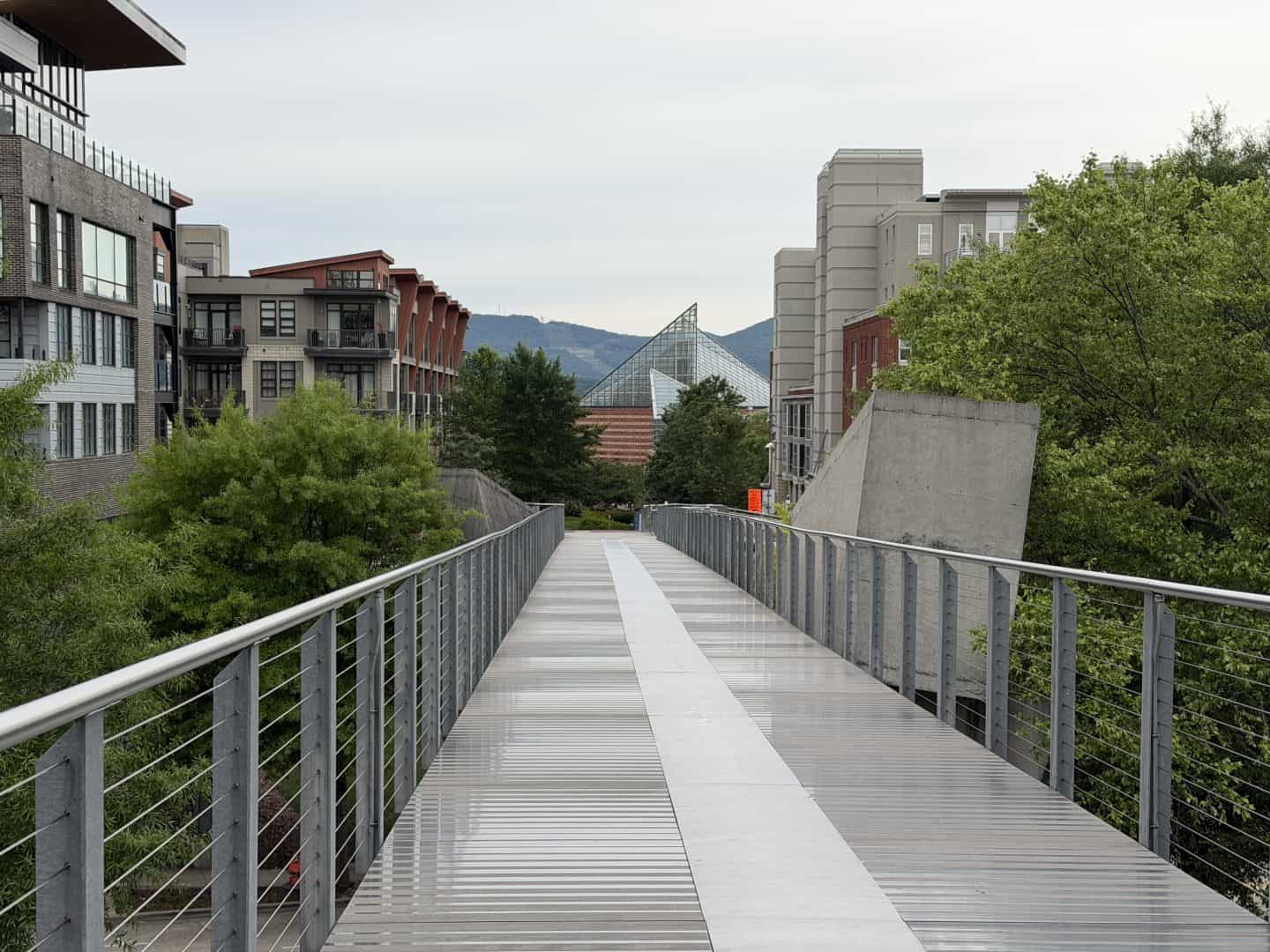 Metal pedestrian bridge lined with rails, leading through an urban campus toward a glass pyramid building, with trees and hills in the background.