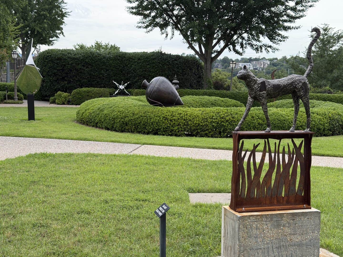 sculpture garden with a bronze cat on a carved pedestal in the foreground with hedges and other sculptures in the background.