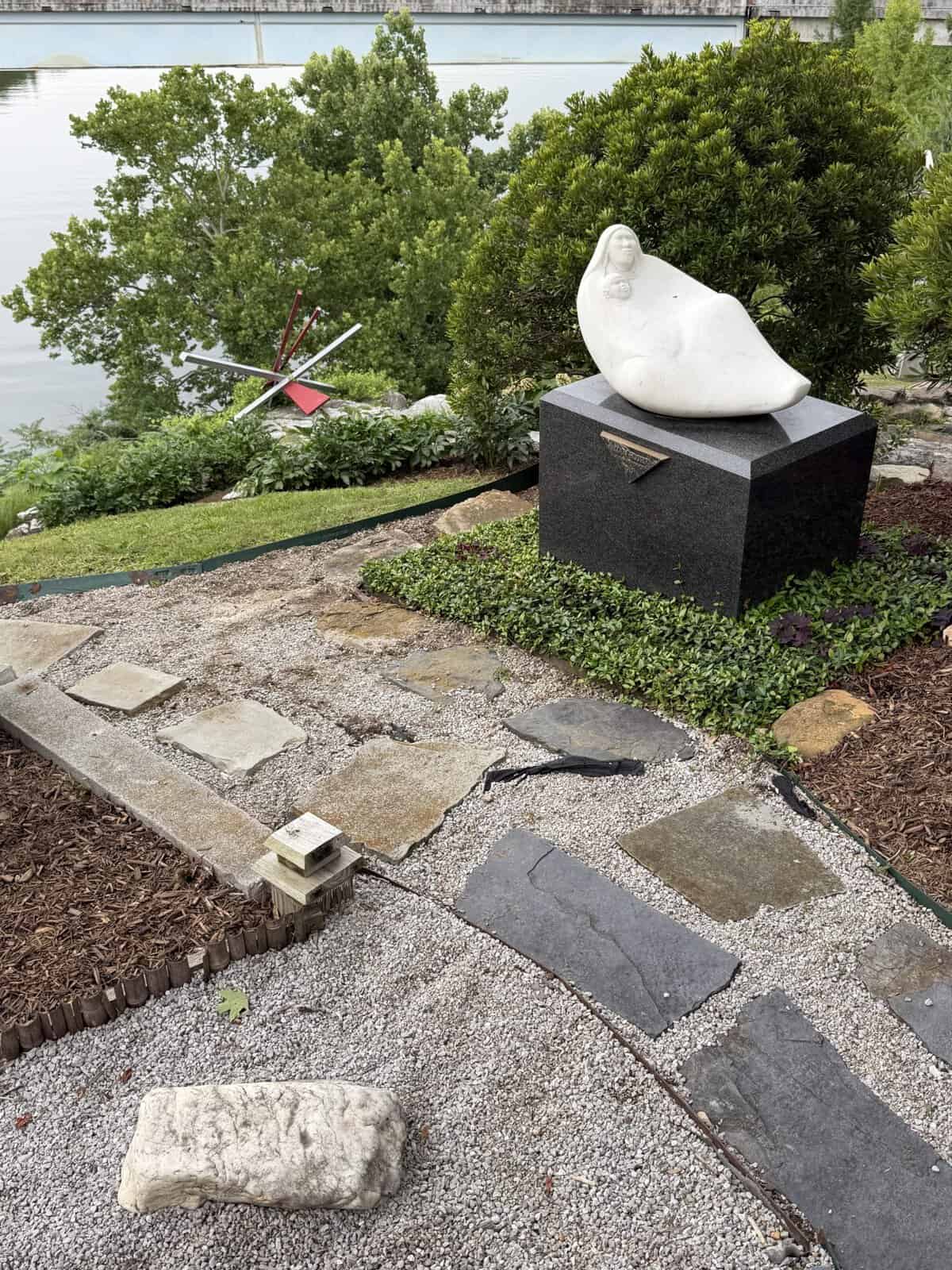Reclining white sculpture on a dark pedestal in a landscaped garden by the water, with green shrubs behind. Alternately, an abstract outdoor sculpture with angular red and white rods near the path.