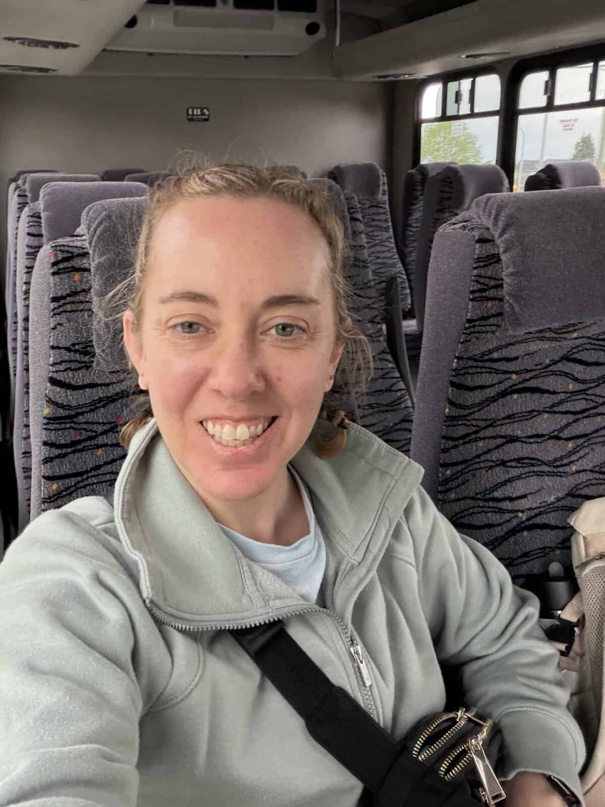 Carli taking a selfie on a bus, wearing a light green sweatshirt while sitting on a charter bus