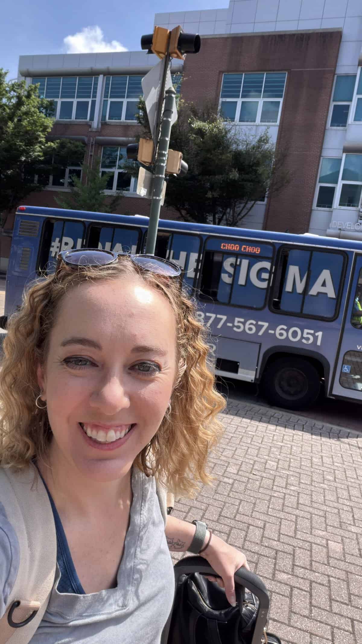 Close-up of a Carli  taking a selfie with chattanooga shuttle bus and a brick building in the background on a sunny day.
