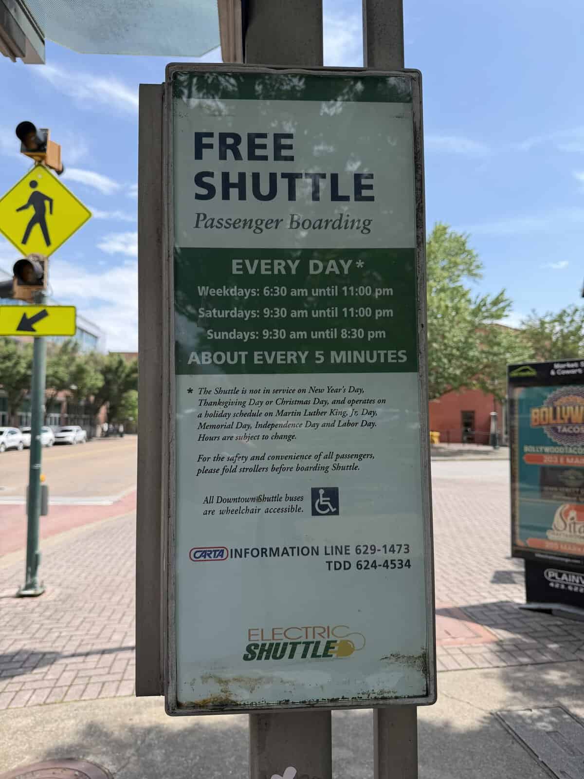 Tall roadside sign advertising a free chattanooga shuttle with operating hours and accessibility note, seen on a sidewalk with a yellow pedestrian crossing sign in the background.