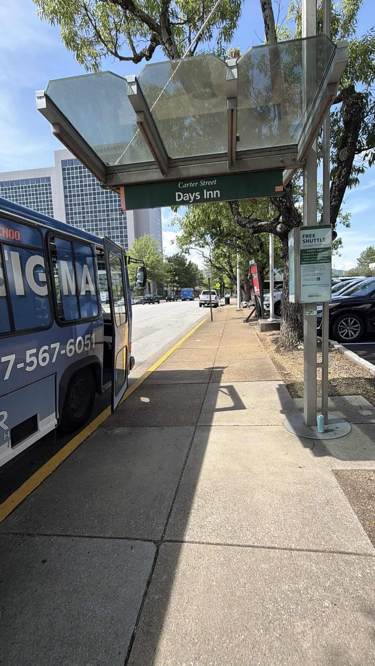 Blue chattanooga shuttle bus stopped at a bus stop beneath a glass canopy with a Days Inn sign overhead on a sunny street.