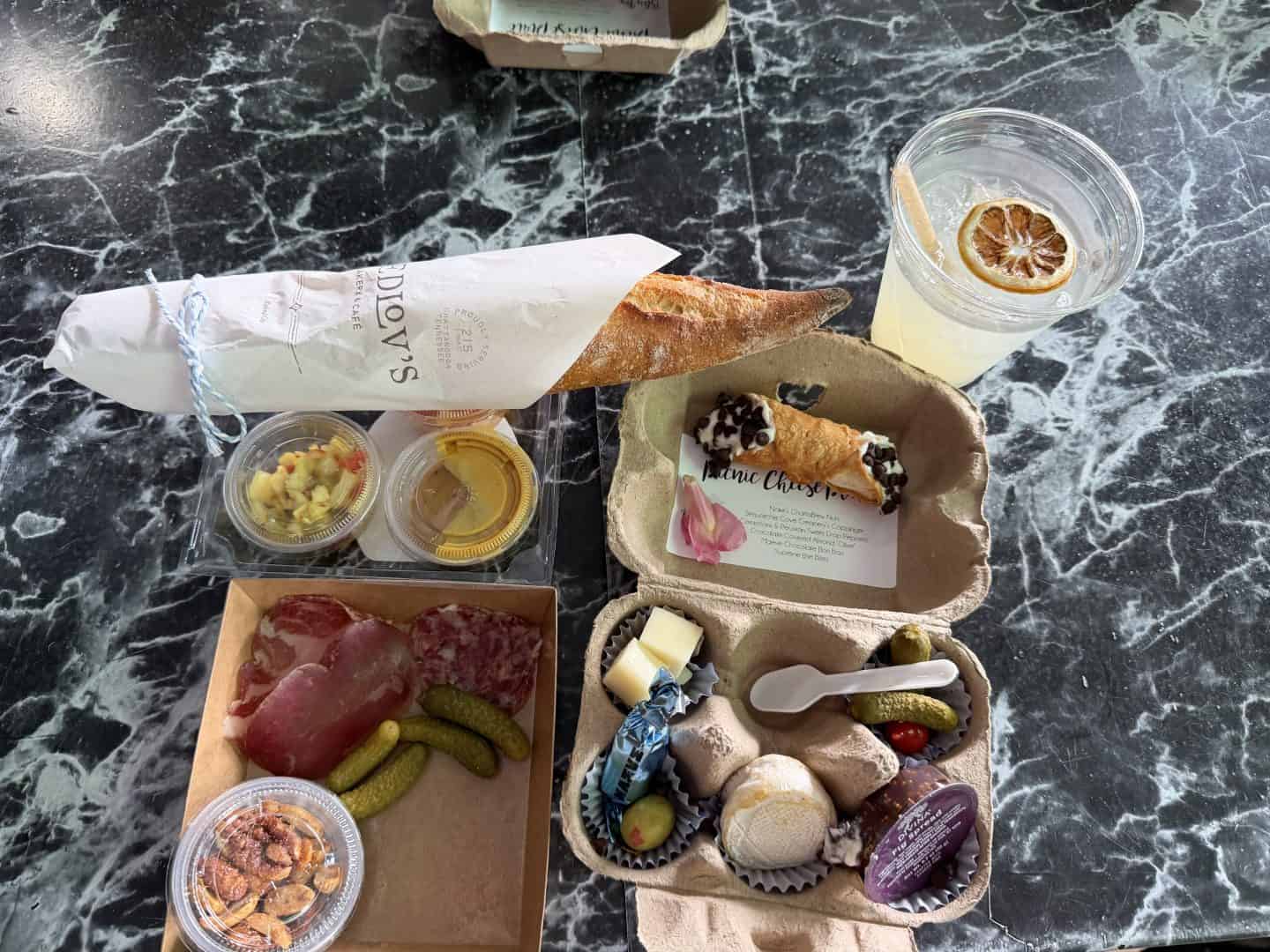 chattanooga food tour picnic items set on a dark marble table: baguette in white wrap, fruit cup, mustard/jam containers, an egg carton with pastries and chocolates, pickles, and cocktail