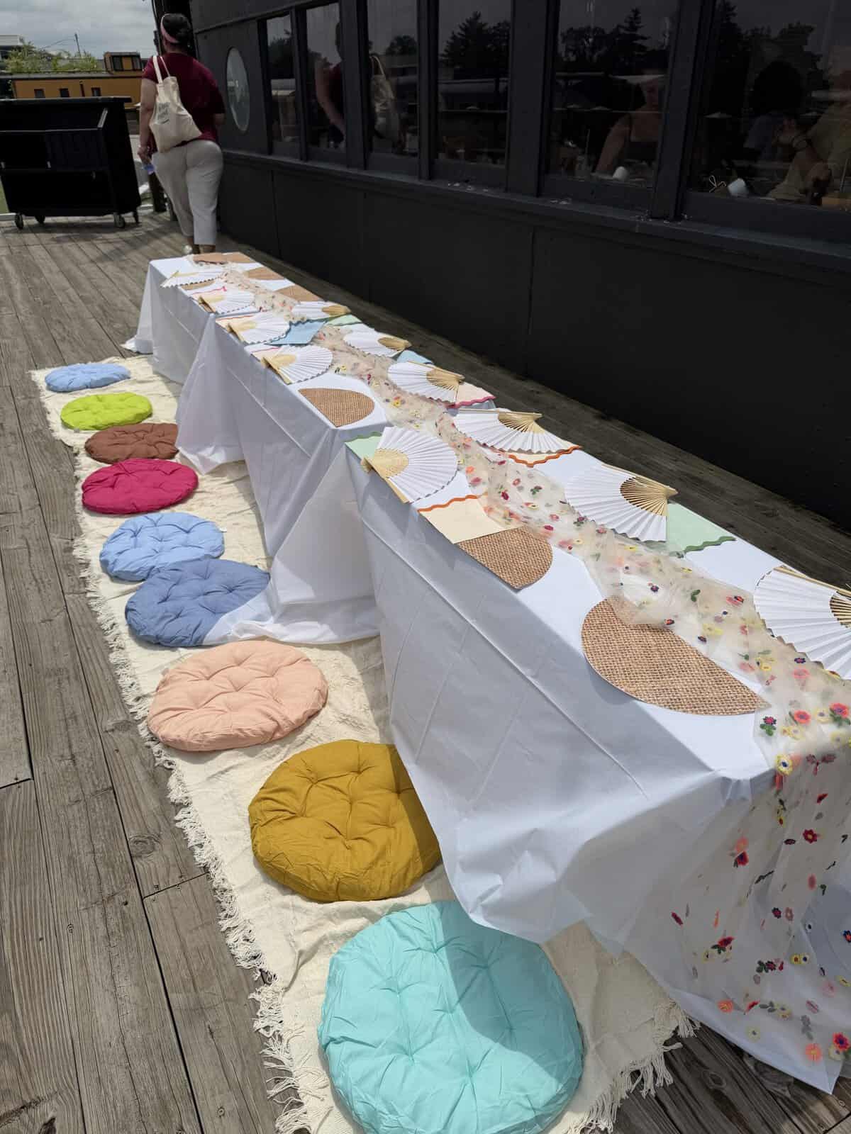 Outdoor event setup: long white-draped table with paper fans and pastel cushions along a beige rug on a wooden deck.