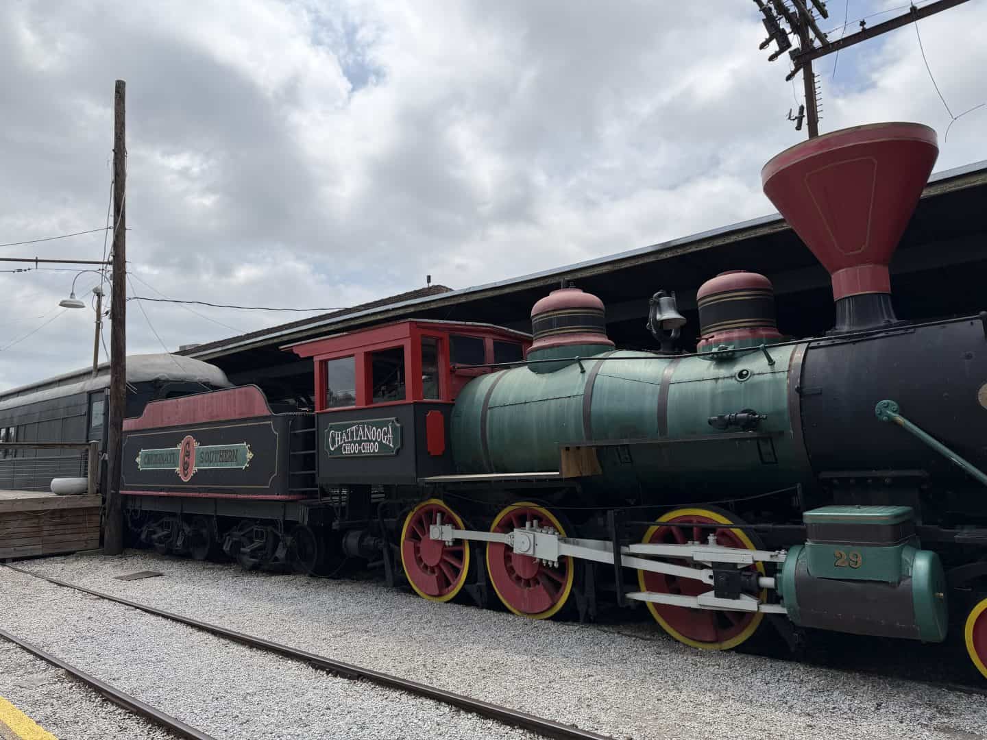 Vintage steam locomotive with red smokestack and green boiler on railroad tracks beside a wooden pole under a cloudy sky.