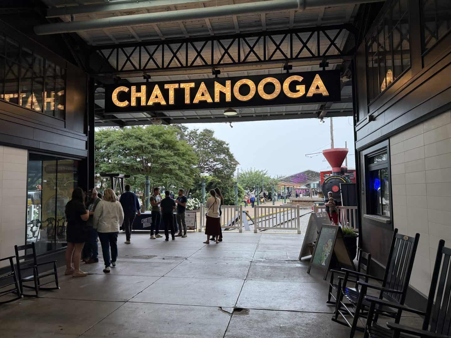 Open-air rail station entrance with a large lit CHATTANOOGA sign overhead and people gathered beneath.