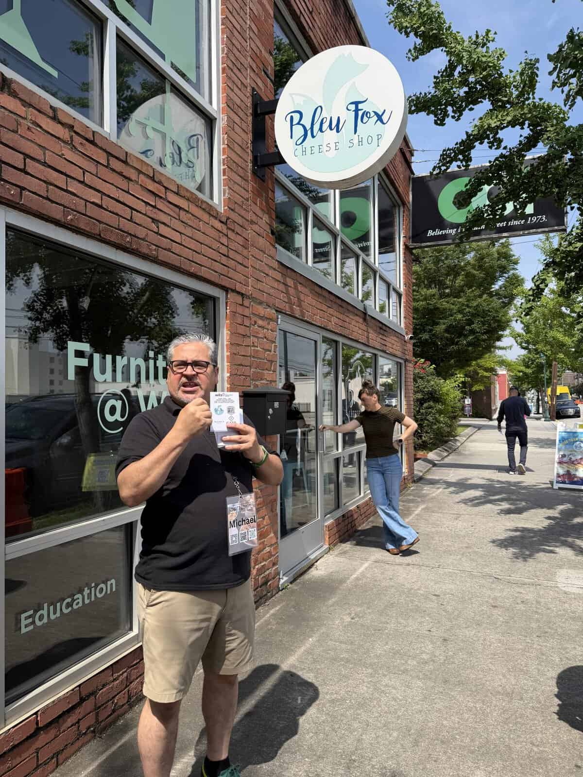 Man with a name badge holding up a flyer outside a brick storefront, Bleu Fox Cheese Shop sign above.