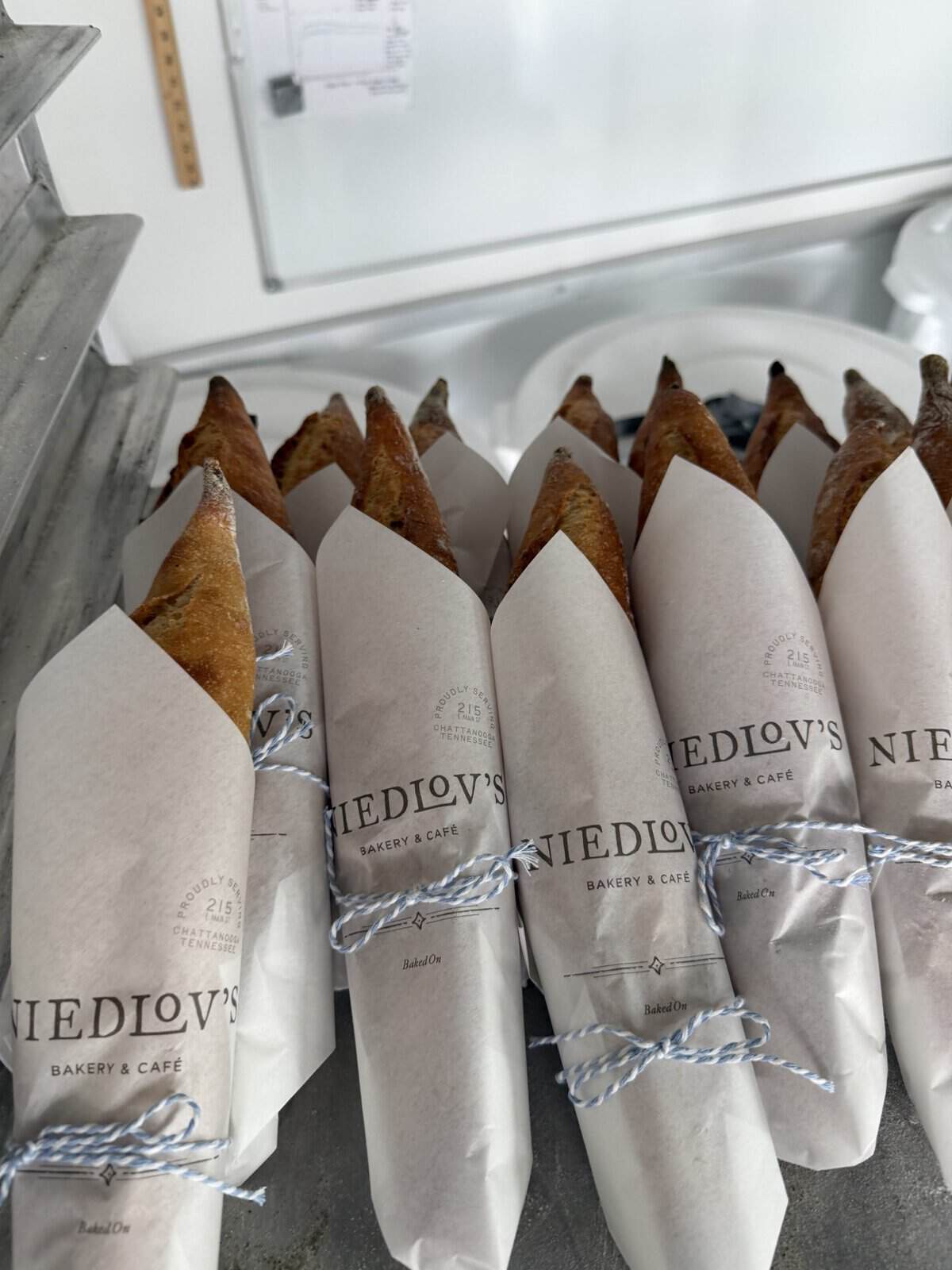 Row of baguettes wrapped in white parchment sleeves with Niedlov's Bakery & Caf&eacute; branding, tied with blue-and-white string, on a bakery display shelf.