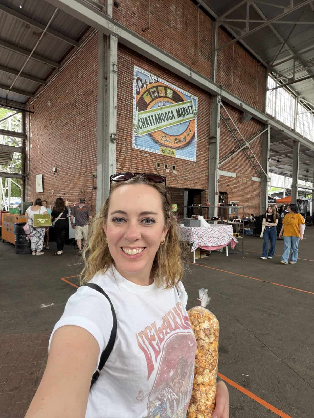 Carli takes a selfie inside a brick-walled market hall with a Chattanooga Market mural in the background and vendors around.