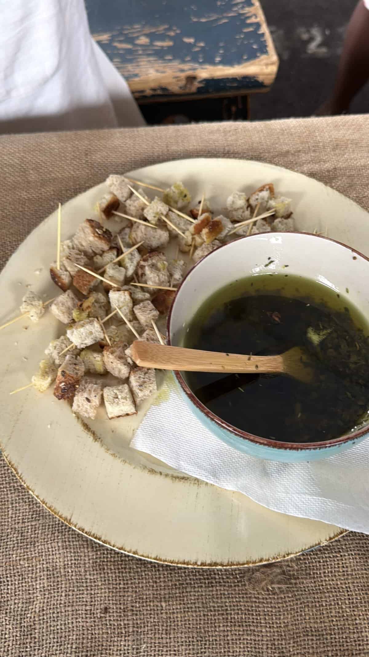 Plate with cubed bread bites on toothpicks surrounding a bowl of green herb infused olive oil on wood table
