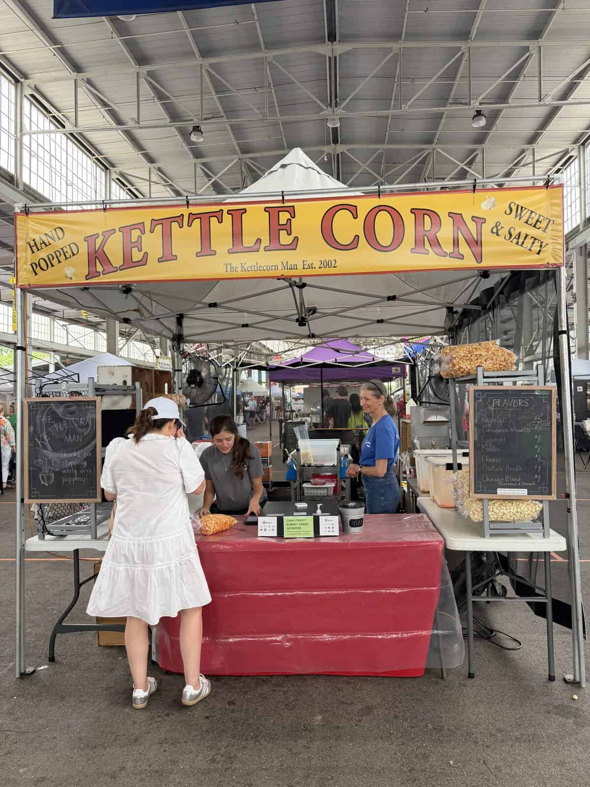 Kettle corn stall at an outdoor market under a metal roof, with a large yellow banner reading'KETTLE CORN' and customers at the counter.