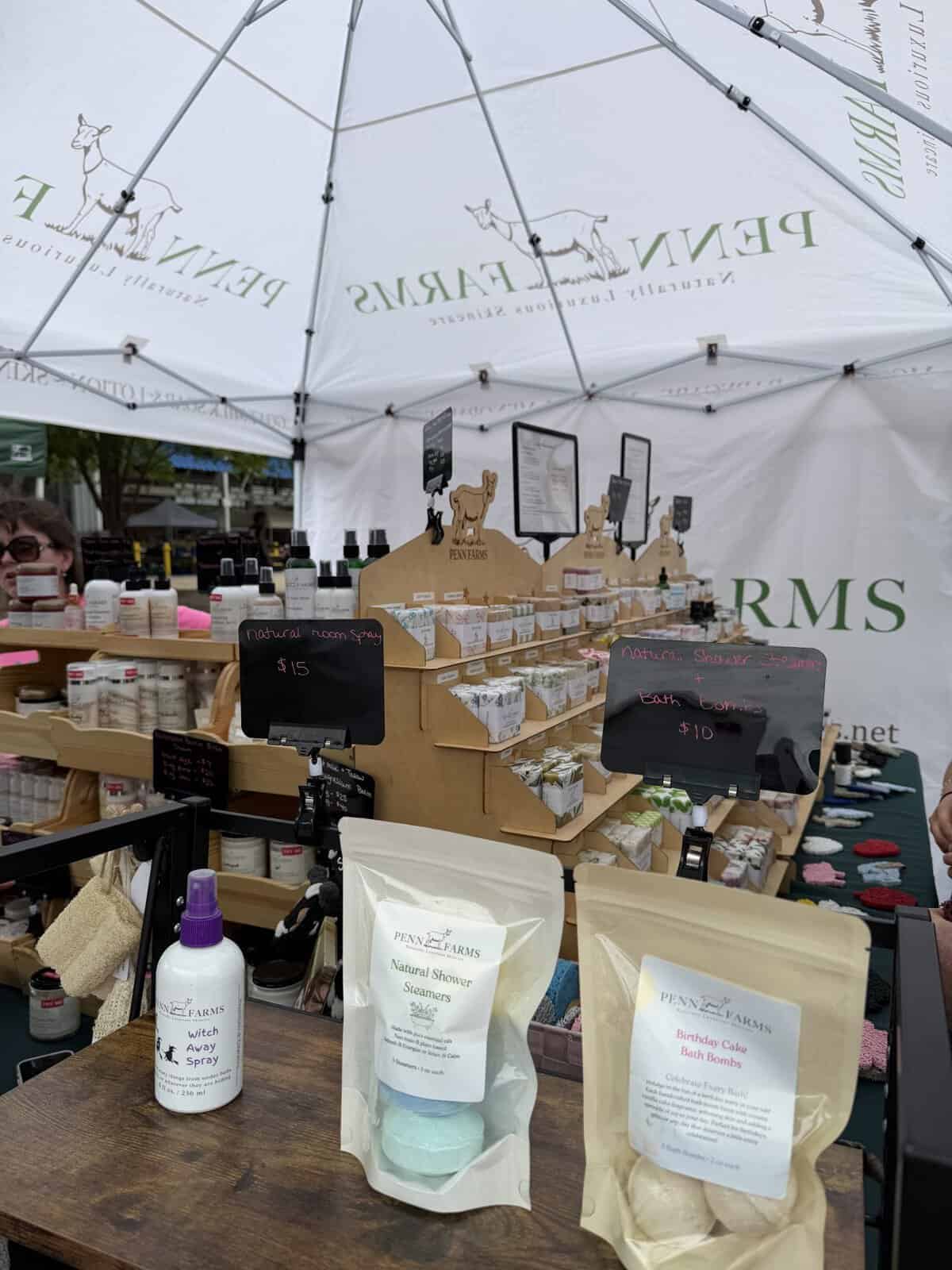 Outdoor market stall selling natural bath products under a white tent with Penn Farms branding; shelves display bottles, bath bombs, and giftsets.