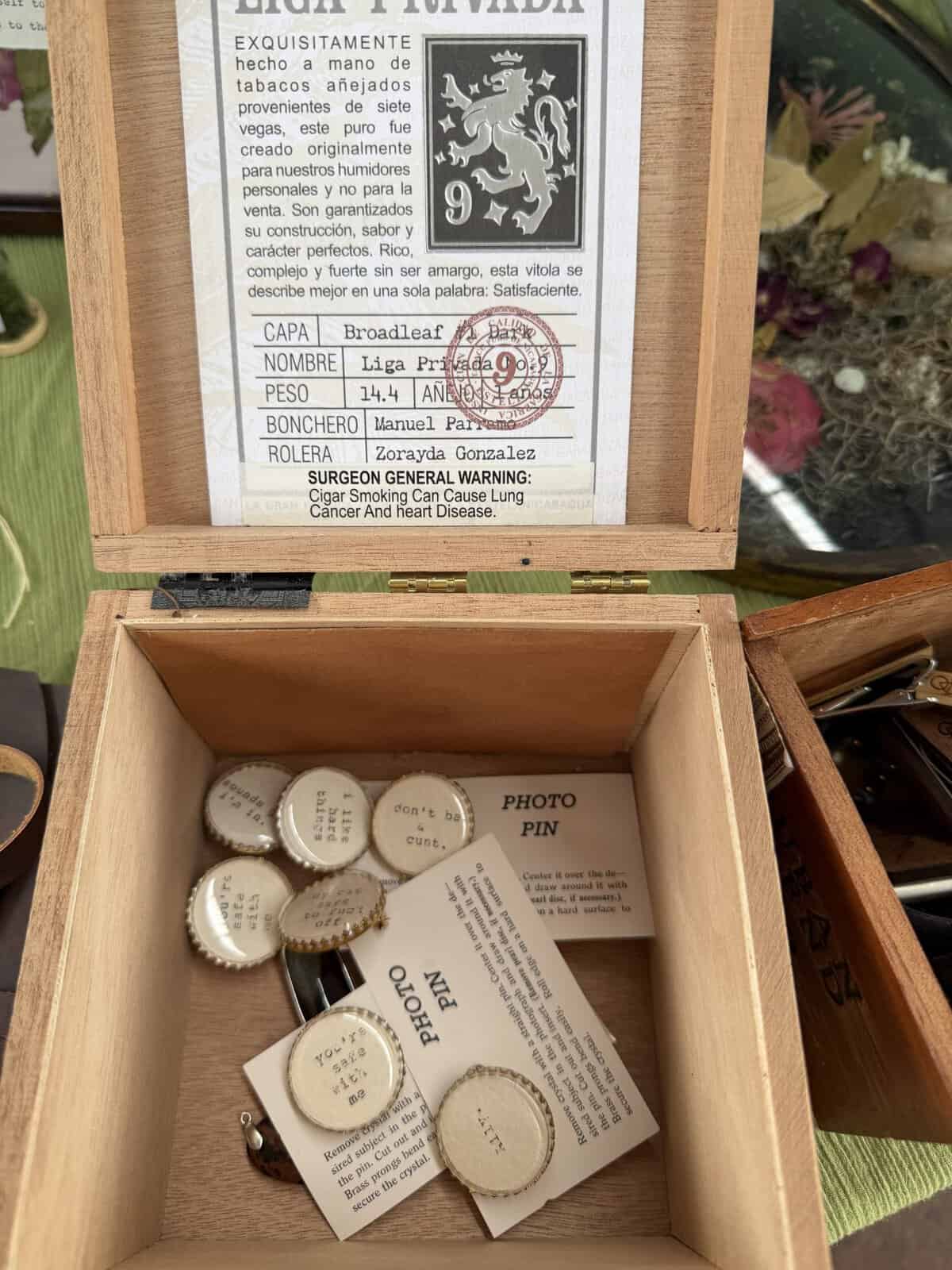 Open wooden humidor with a printed label featuring a lion crest and form fields (CAPA, NOMBRE, PESO) and round caps visible in the lower tray.