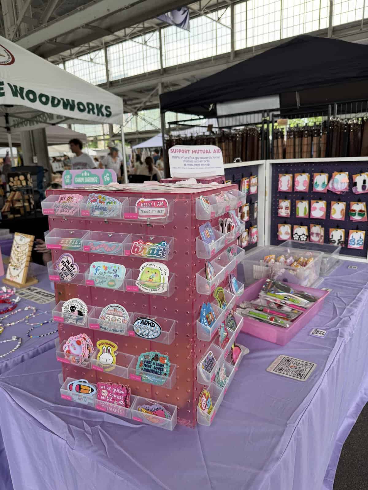 Pink pegboard display filled with colorful badges and pins at a craft fair booth, purple tablecloth underneath and other vendor tables in the background.