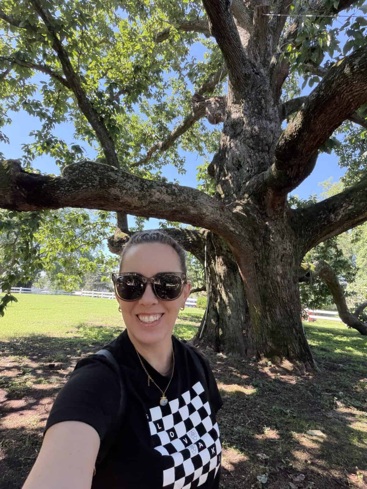 Carli in black shirt & pink skirt taking a selfie under magnolia tree with large winding branches