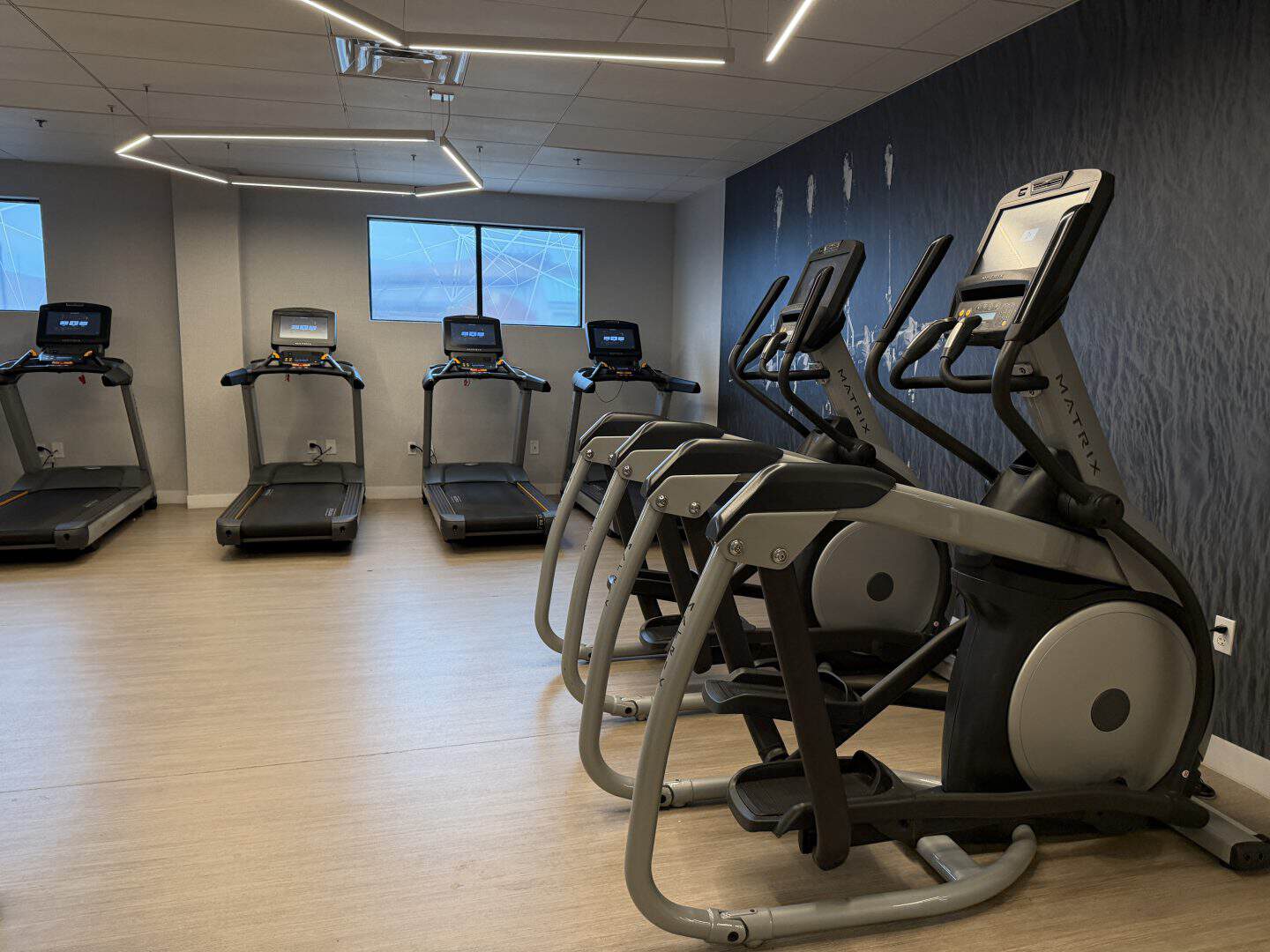 Row of treadmills in a modern gym with a dark accent wall and geometric ceiling lights above the machines.
