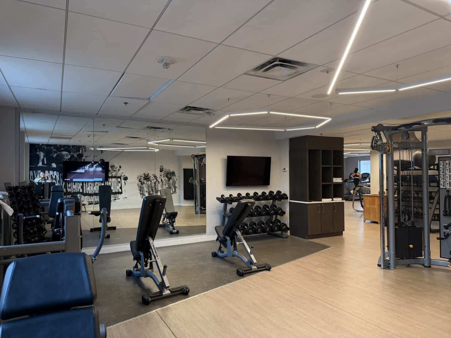 Spacious gym interior with cardio machines, free weights, mirrors along the wall, and modern ceiling lights.