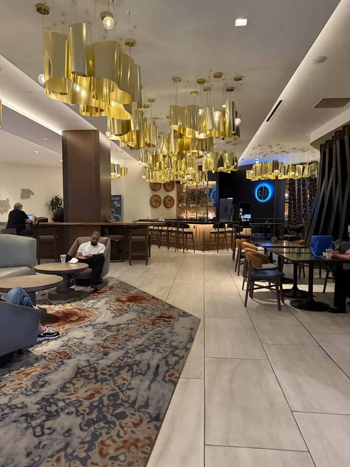 Luxurious hotel bar area with cascading gold pendant lights and a long wooden bar, seating, and a patterned rug.