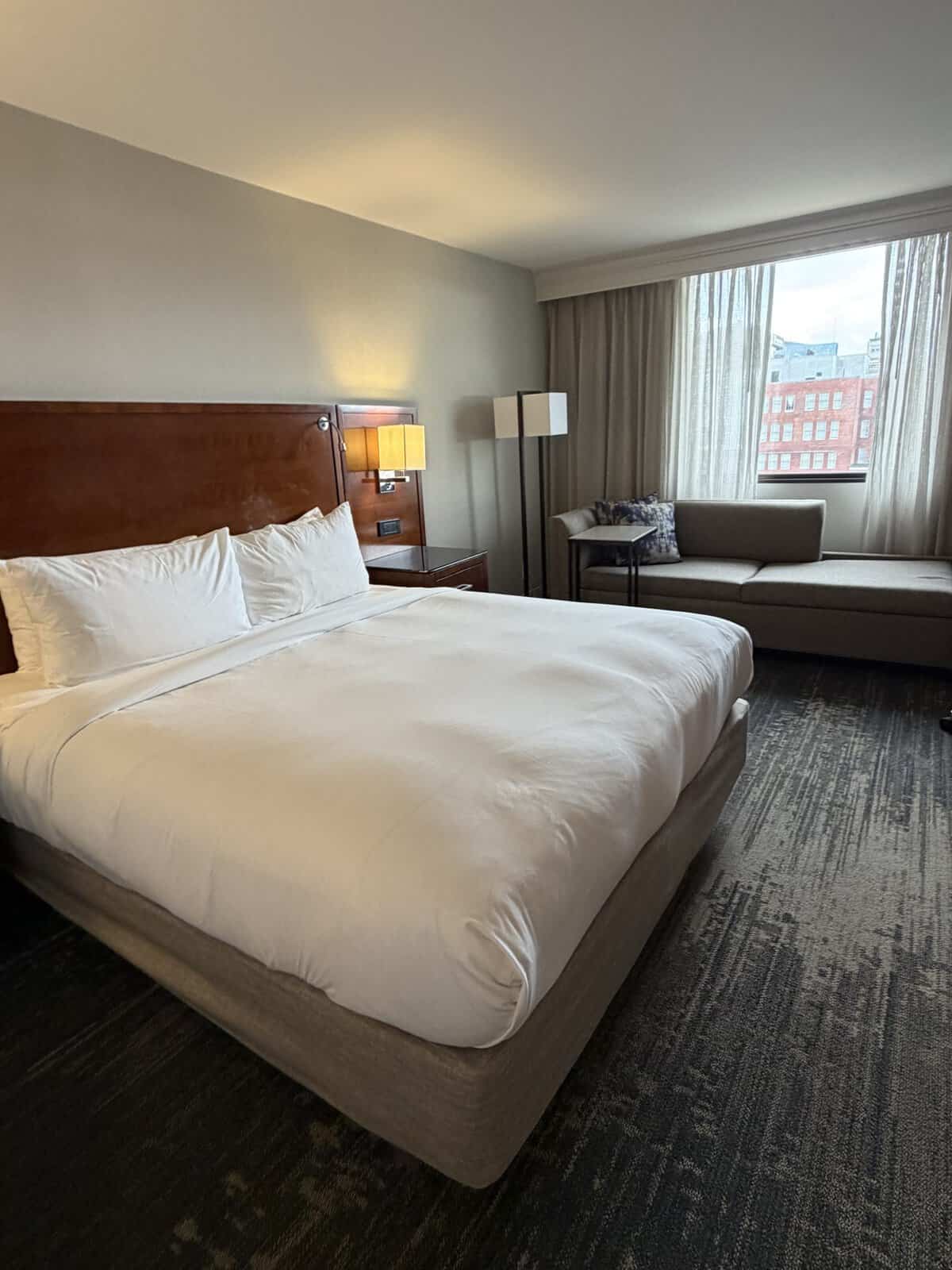 Hotel room featuring a large white-bleed bed with a dark wood headboard and bedside lamps, plus a beige sofa by a window with sheer curtains and a small table nearby; city buildings visible outside the window.