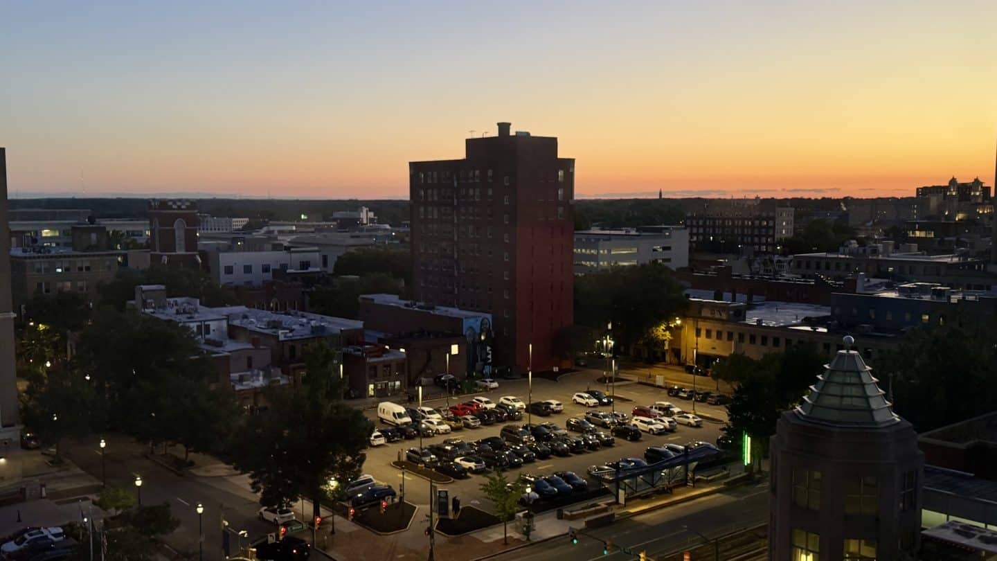 Richmond skyline at sunset viewed from Downtown Richmond Marriott, with a tall brick building and a glowing orange horizon over a parking lot and streets below.