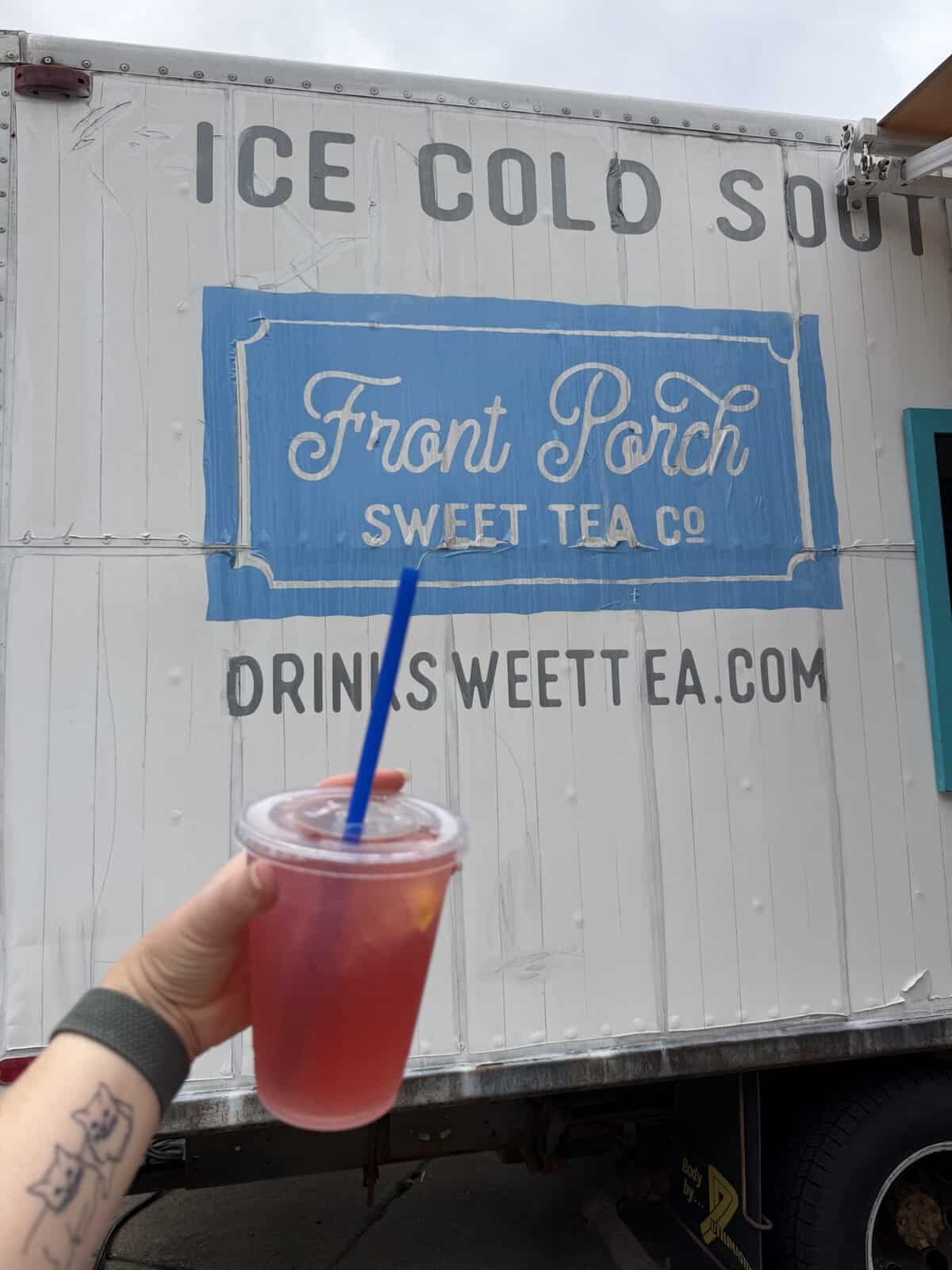 Person holding a cold red tea or beverage with a blue straw in front of a white trailer sign reading'Front Porch Sweet Tea Co' and a web address below.