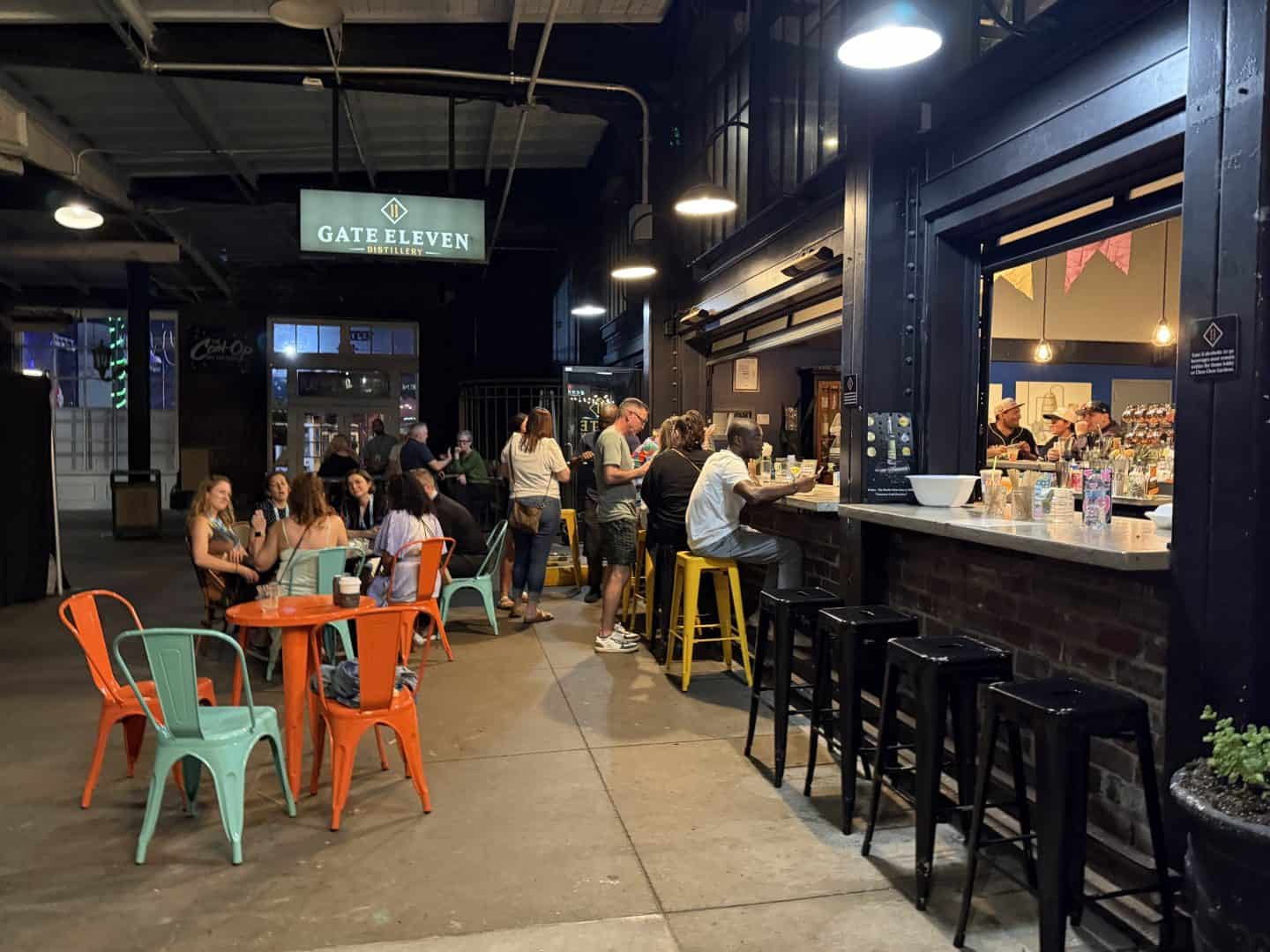 exterior seating of gate eleven distillery iwth people congregated at bar window.  orange and teal tables and chairs in foreground with group of women at a table behind